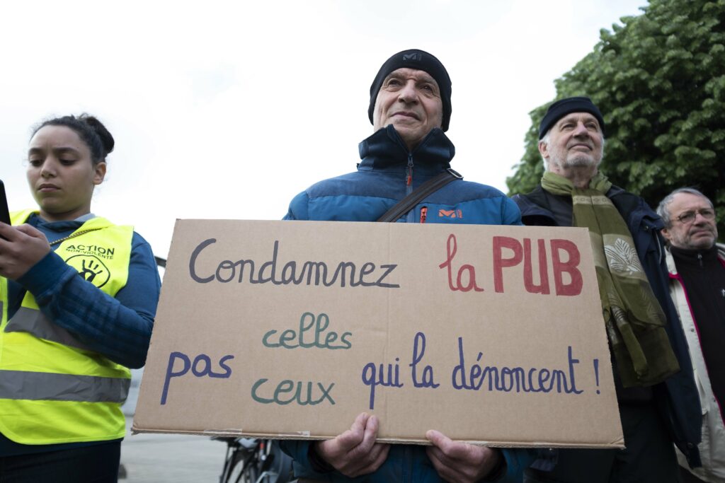 Gros plans sur une pancarte que tient un militant, où il est écrit " Condamnez la pub, pas celle et ceux qui la dénoncent!"