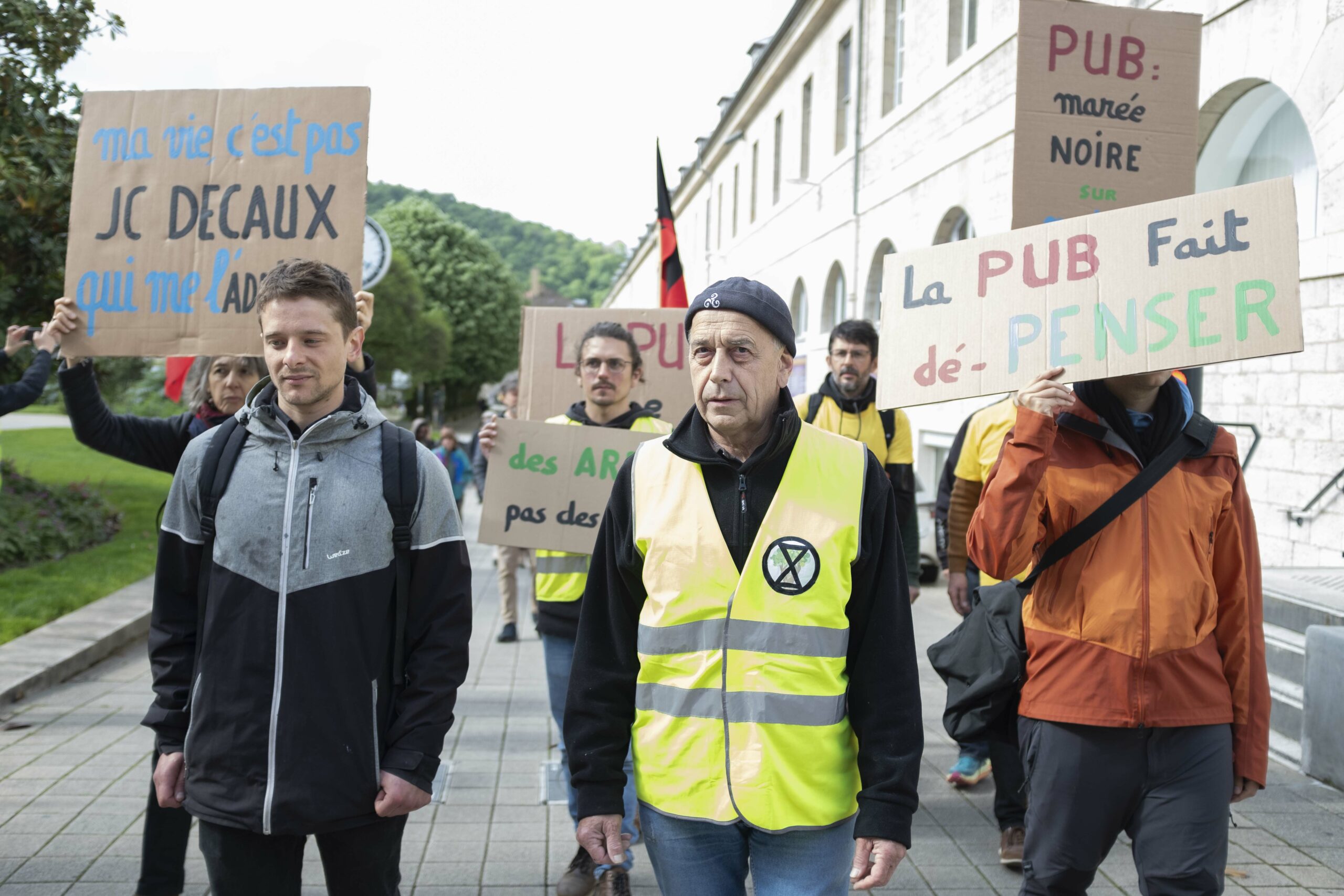 Sur le parvis de la mairie, des militants avancent vers le tribunal. A gauche au premier plan on retrouve Tristan, au centre un militant au chasuble jaune XR et a droit un militant portant une pancarte "La pub fait dépenser".