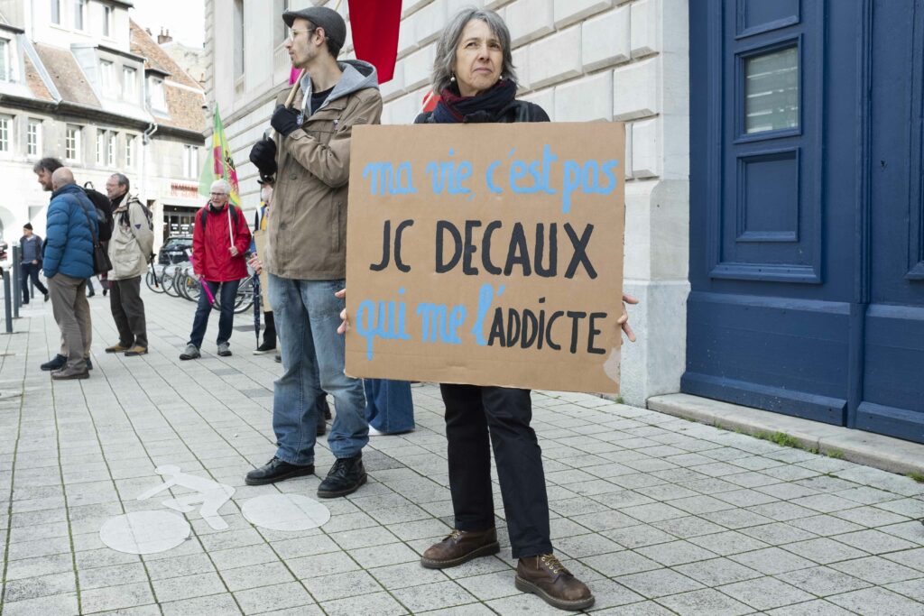 Devant les mur du tribunal, une militante tient une pancarte où il est écrit "ma vie, c'est pas JC Decaux qui me l'addicte.