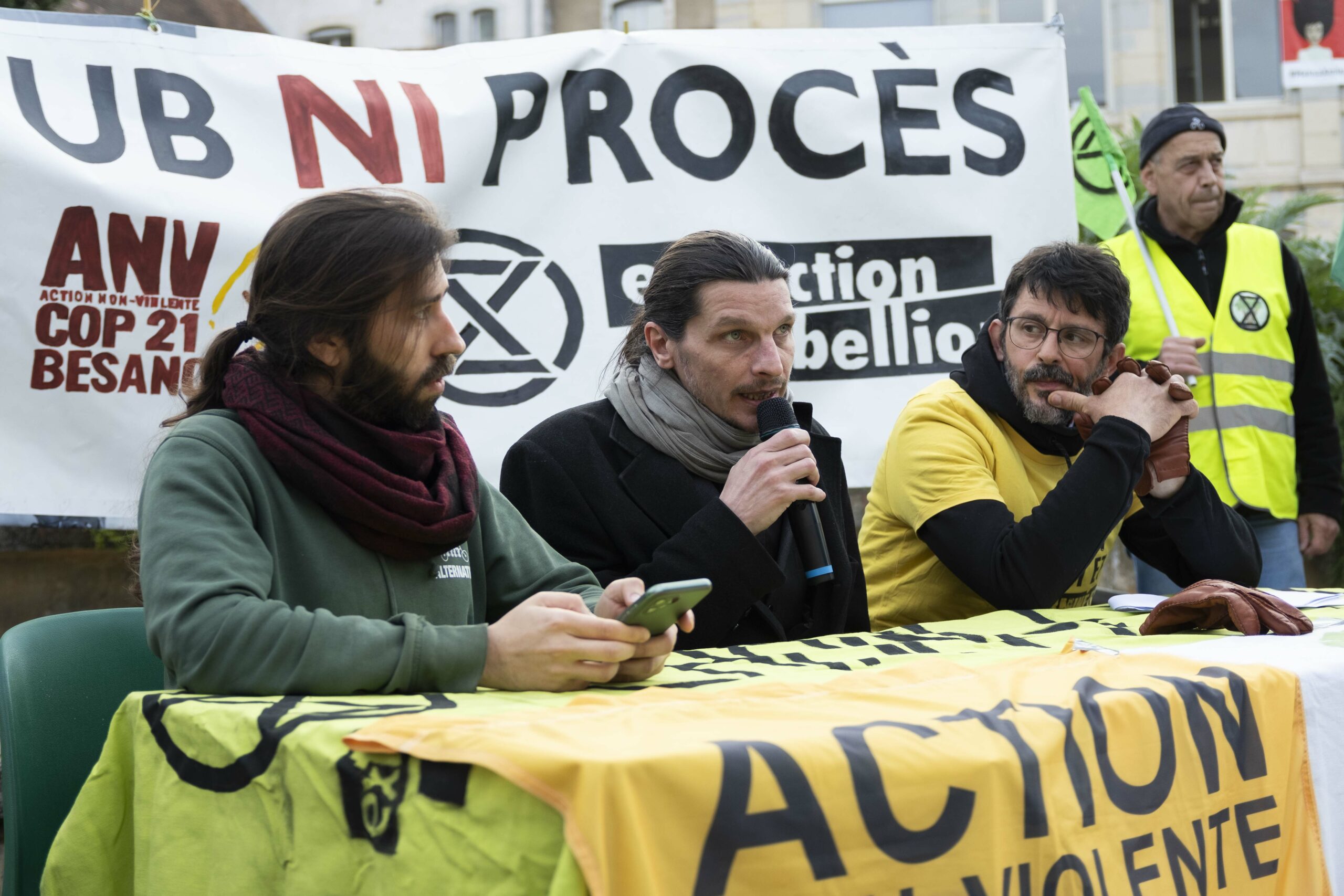 Autour d'une table installée sur le parvis de la mairie de Besançon, Thomas Bourgenot, chargé de plaidoyer pour l'association Résistance à l'Agression Publicitaire prend la parole. Il est entouré du porte parole d'Alternatiba et d'un militant d'ANV Cop 21.