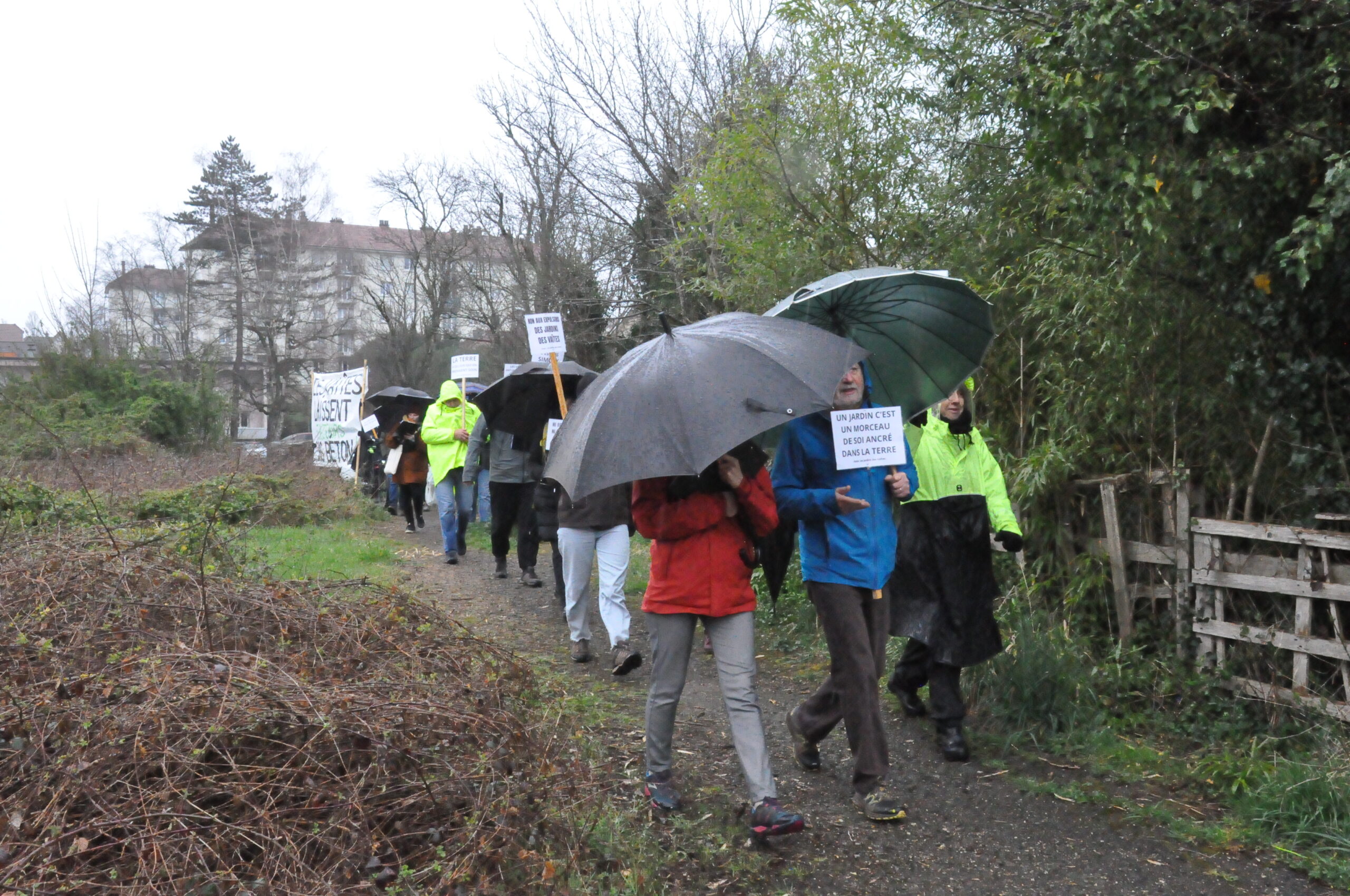 Quelques manifestant·es, abrités sous des parapluie, sur un chemin mouillé, avec leurs pancartes