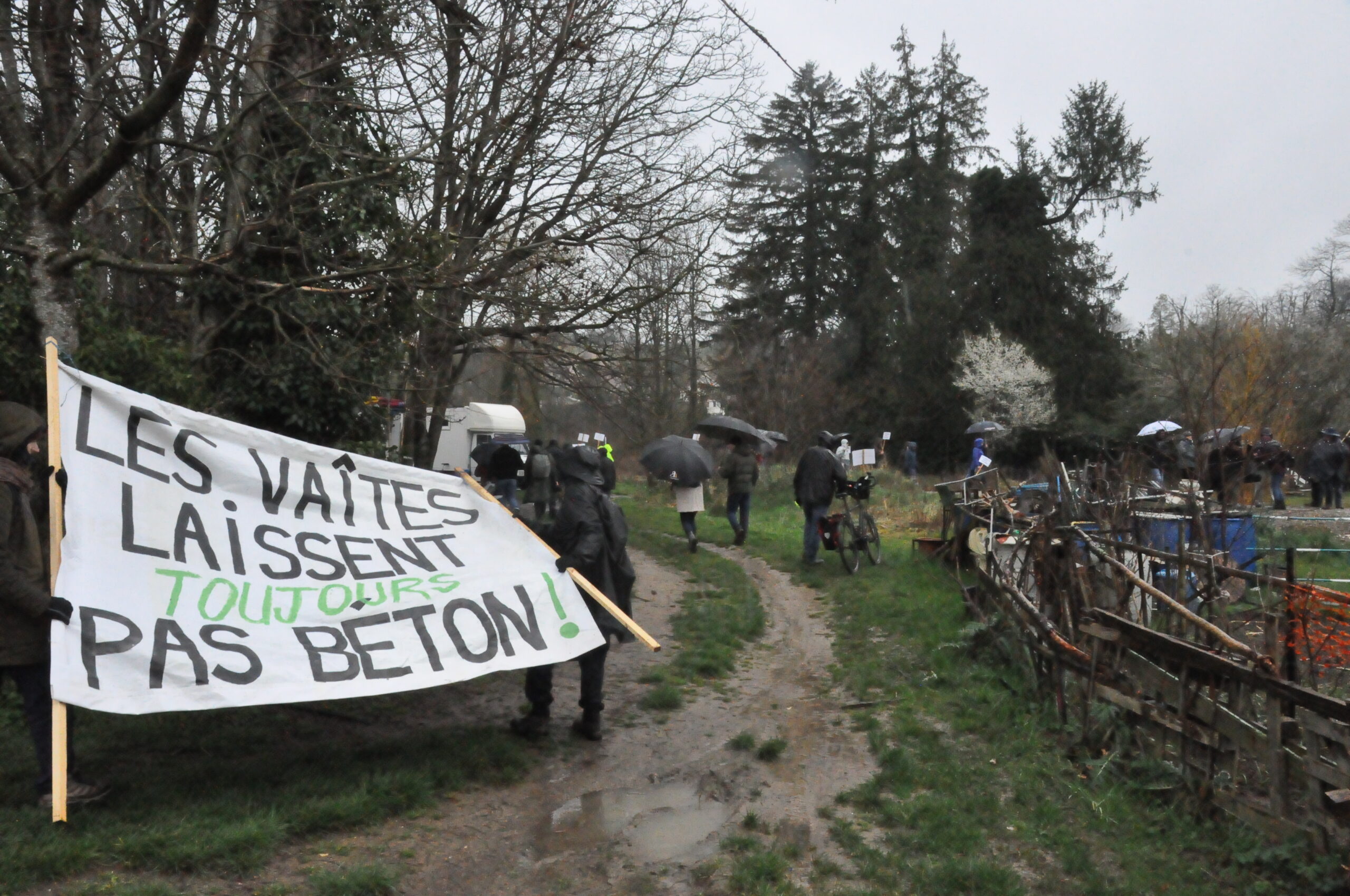 Quelques manifestant·es sur un chemin mouillé, en habits de pluie, avec une pancarte "les vaites laissent toujours pas béton"