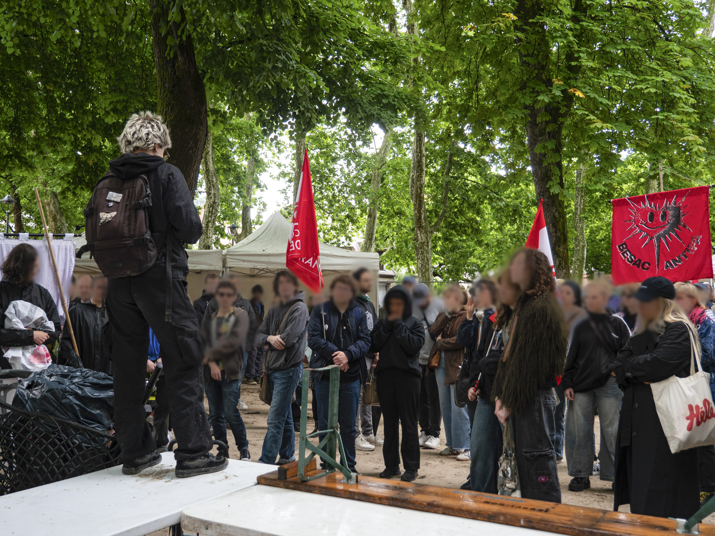 Debout sur une table, une personne s'adresse à une foule captivée, place Granvelle à Besançon. On aperçoit un drapeau Besac Antifa et les stands en arrière plan