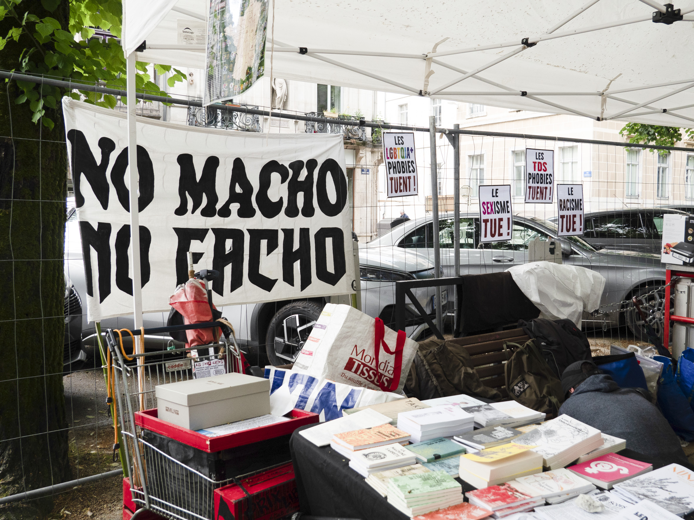 Une énorme banderole No macho no facho affichée sur une grille, devant un stand couvert de livres. On peut également y lire des affichettes les LGBTQIA+ phobies tuent, le sexisme tue, les TDS phobies tuent, le racisme tue