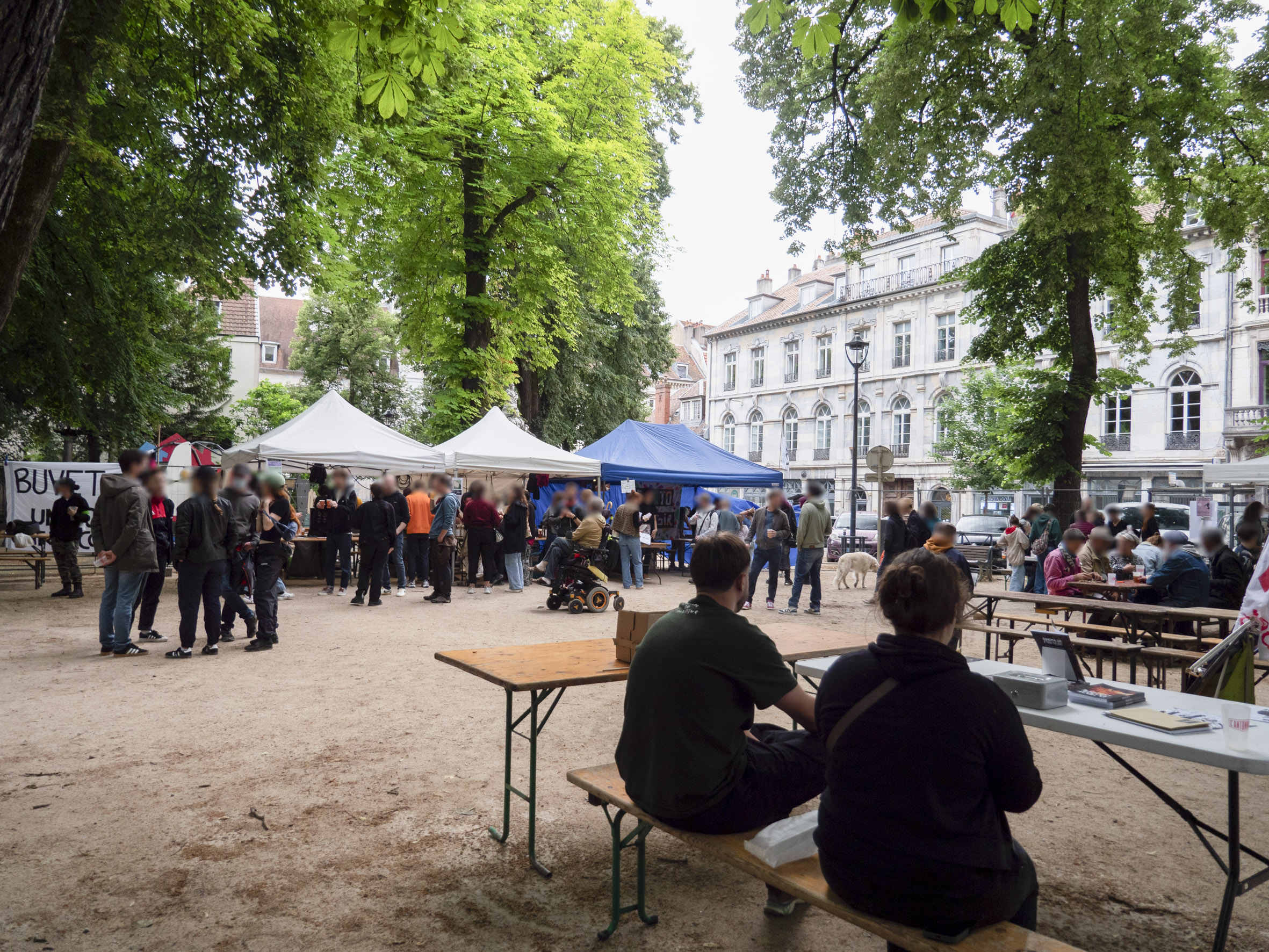vue d'ensemble du festival, place Granvelle, avec divers stands et tables, où des gens discutent un peu partout, assis ou debout, sous les arbres
