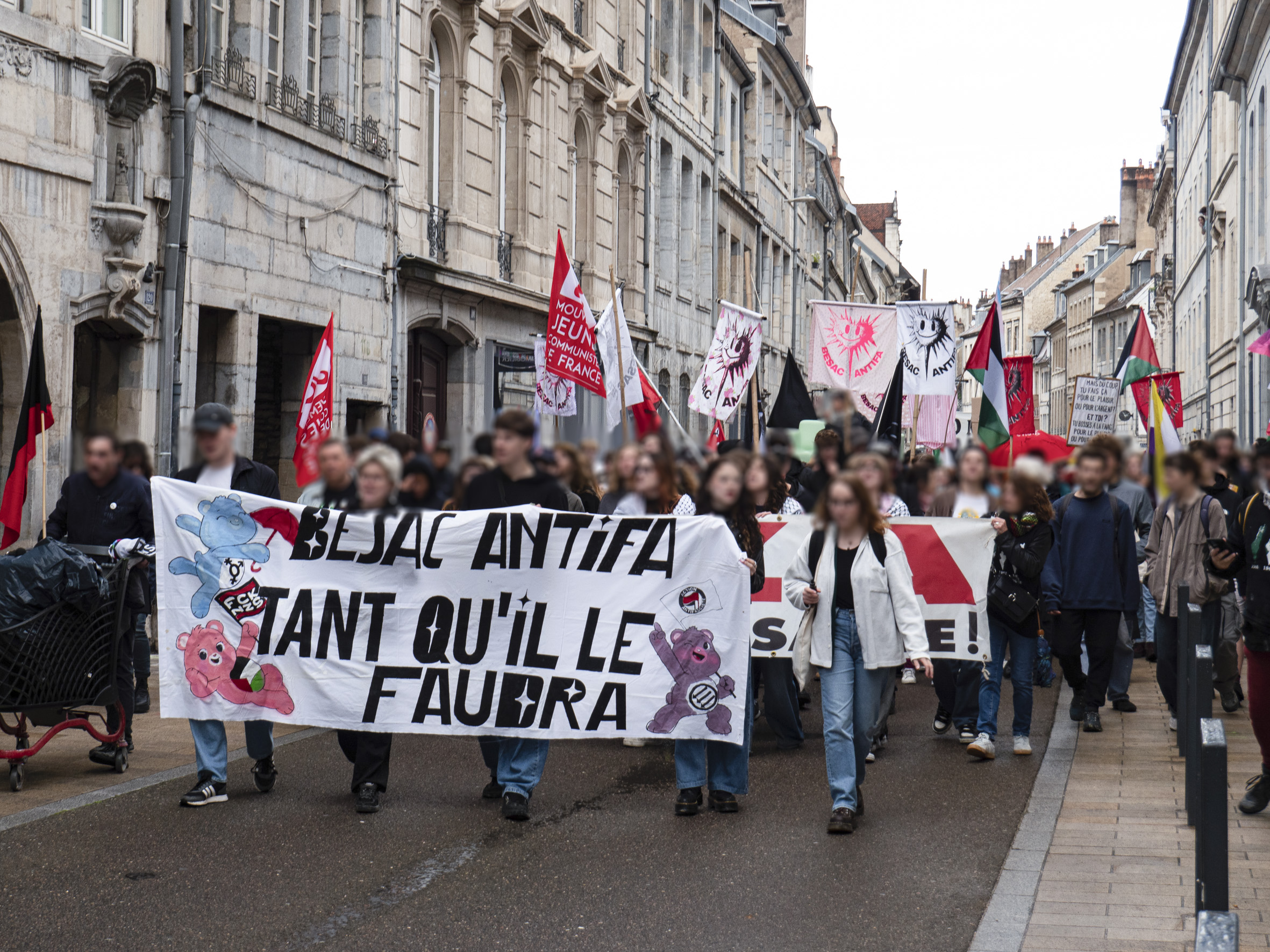 Manifestation dans les rues de Besançon (grande rue), avec en tête de cortège la banderole Besac Antifa tant qu'il faudra décorée de Bisounours.
De nombreux drapeaux sont visibles : palestinien, trans, communiste, anarchiste, antifasciste