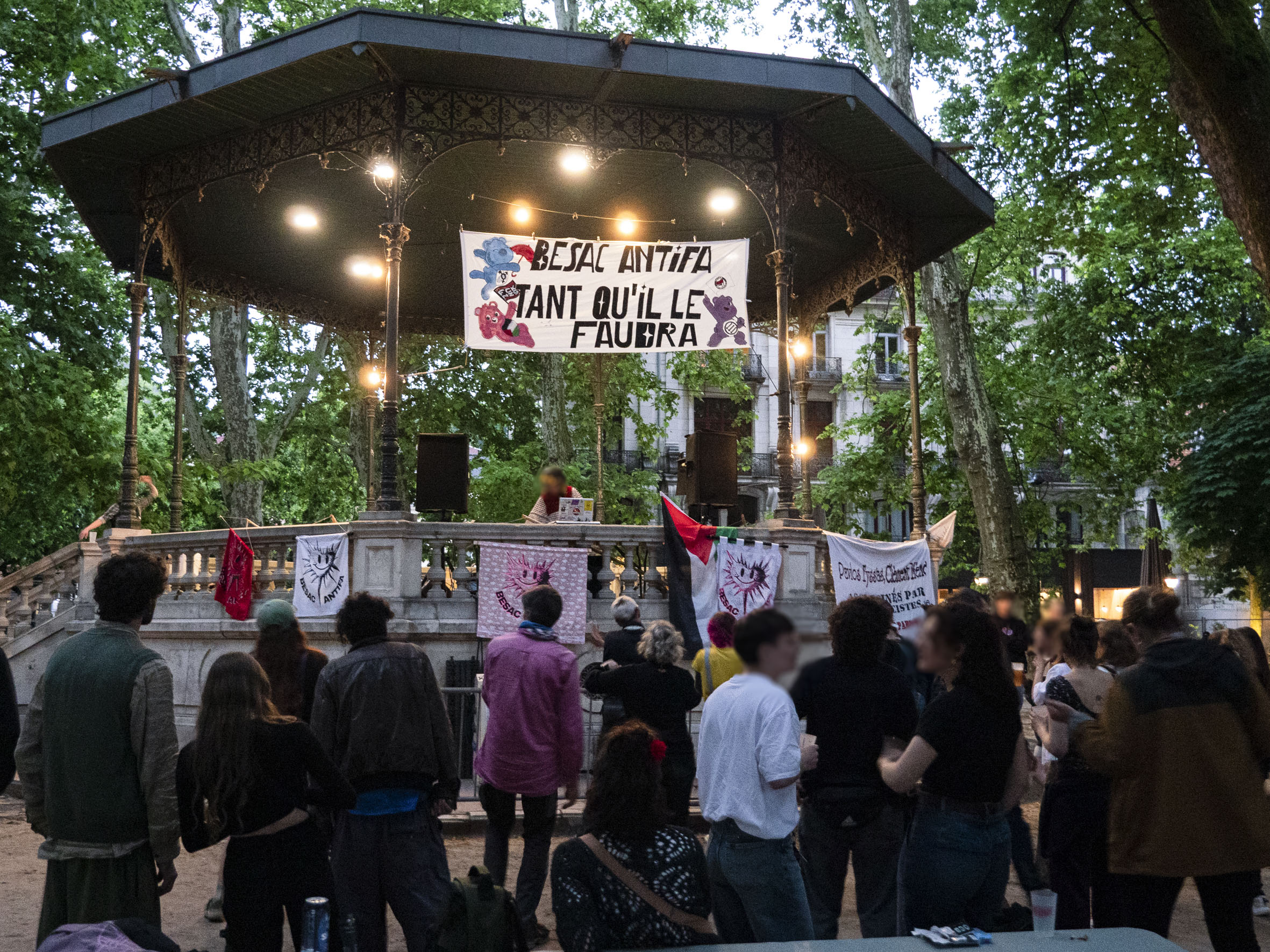 la banderole "Besac antifa tant qu'il faudra" accrochée dans le kiosque de Granvelle et bien éclairée, alors que la lumière du jour commence à décliner. Les gens dansent au son de la musique