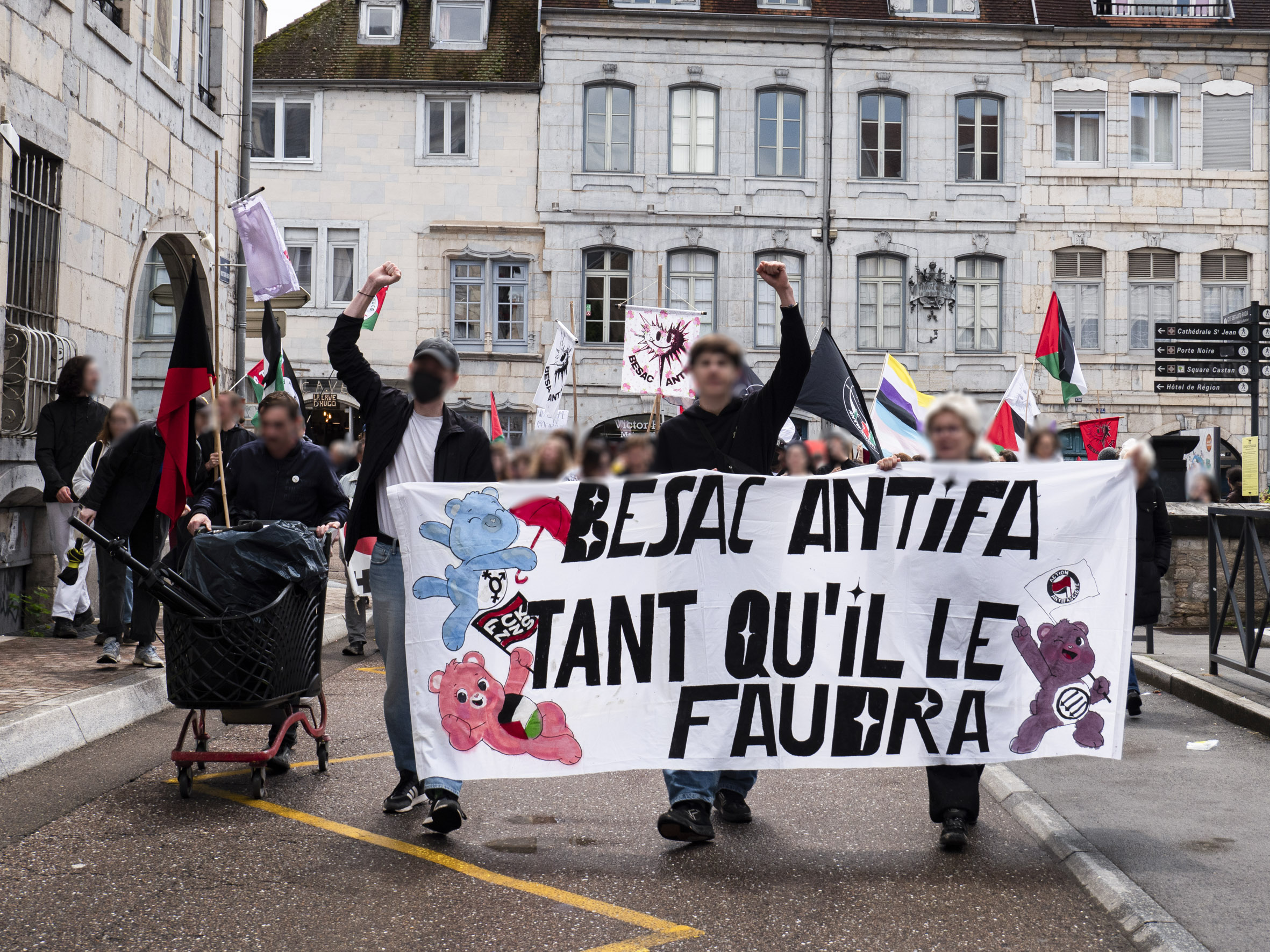 Manifestation dans les rues de Besançon, avec en tête de cortège la banderole Besac Antifa tant qu'il faudra décorée de Bisounours.
De nombreux drapeaux sont visibles : palestinien, trans, communiste, anarchiste, antifasciste, ainsi qu'un sound system transporté dans un caddie