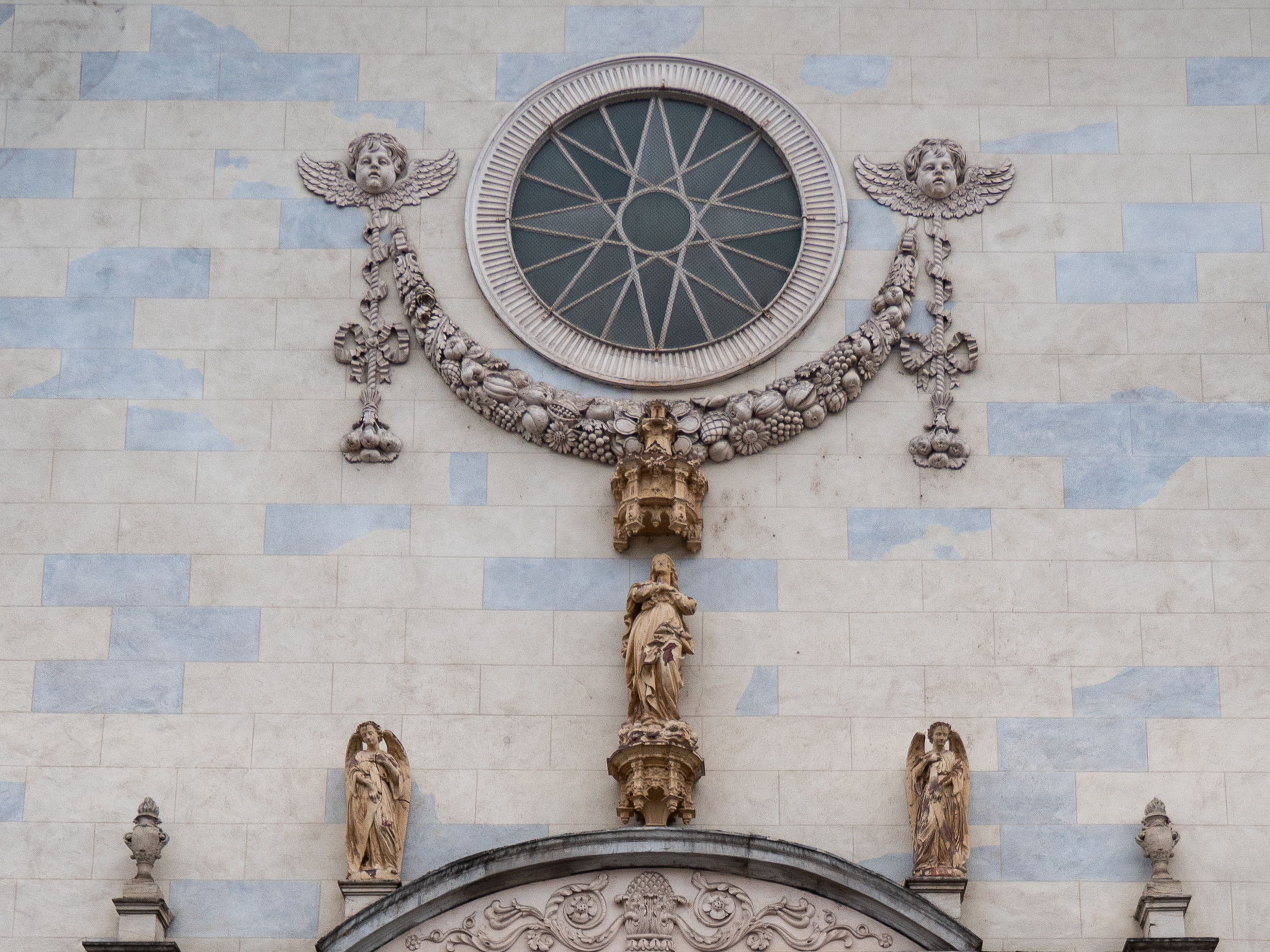 rosace d'église avec une étoile à huit branches, entourée de deux têtes d'angelots montée sur caducées ailés, reliés entre eux par une écharpe de fruits et de fleurs, le tout surplombant quelques sculptures au-dessus d'un fronton de porte que l'on devine au bas de l'image