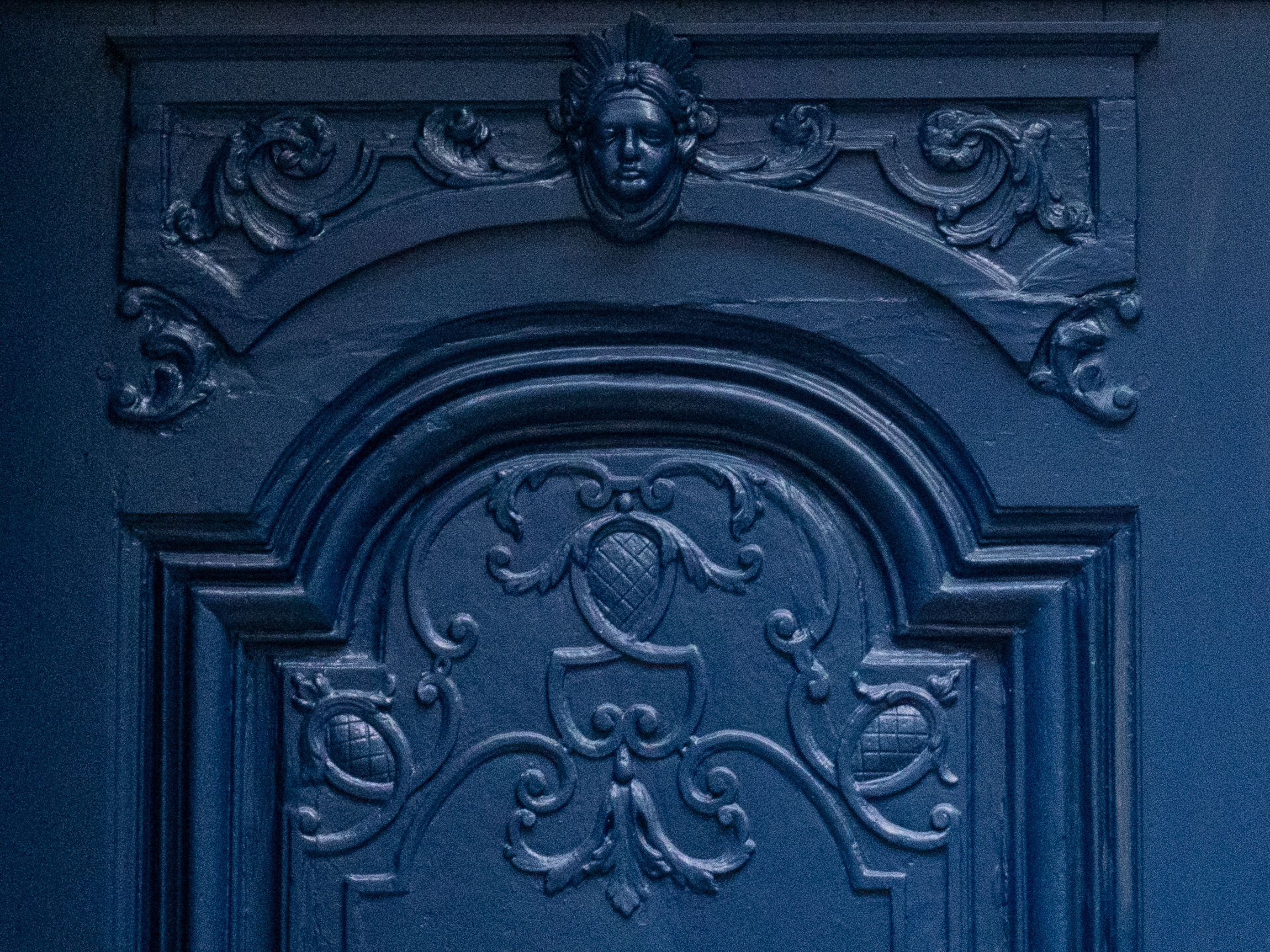 sculpture en bas relief dans un panneau en bois, représentant une tête dans sa partie haute, avec en dessous des motifs floraux, le tout peint en bleu sombre