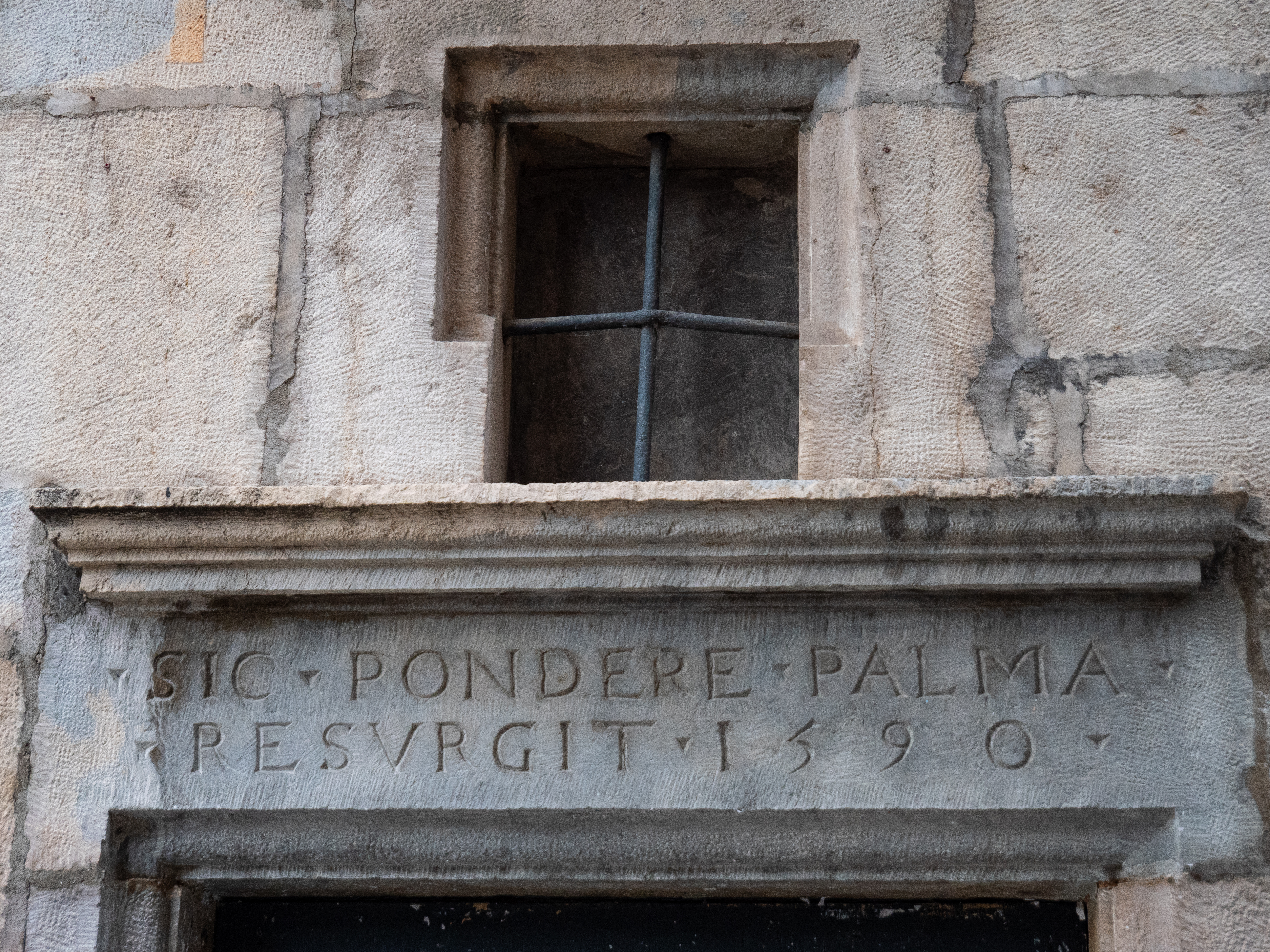 fenêtre constituée de deux barreaux perpendiculaires dans une ouverture en pierre, au dessus d'un fronton sur lequel on peut lire SIC PONDERE PALMA RESURGIT 1590