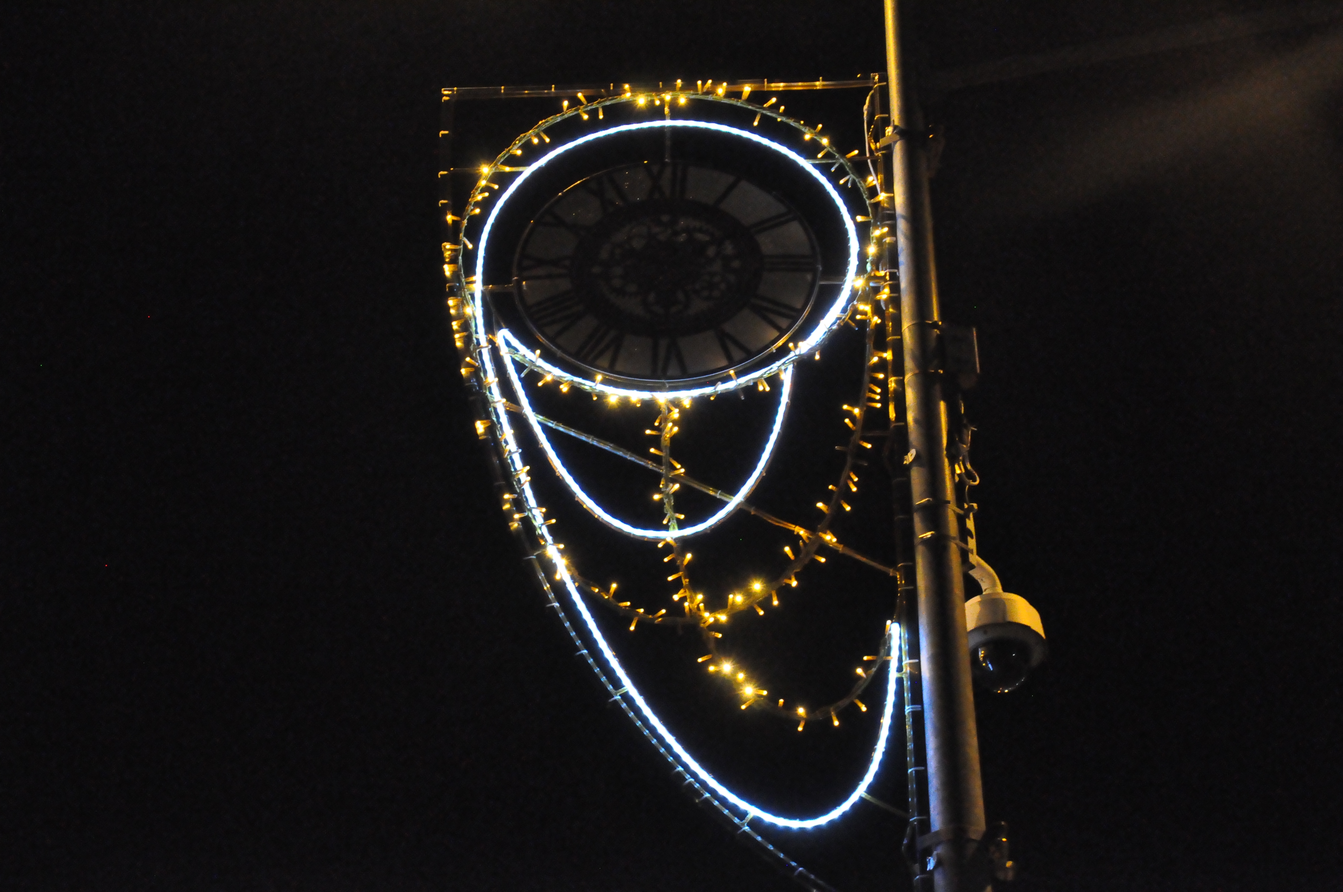 Gros plan d'une illumination en horloge (toujours pas éclairée) sur un réverbère, également affublé d'une caméra de vidéosurveillance. 