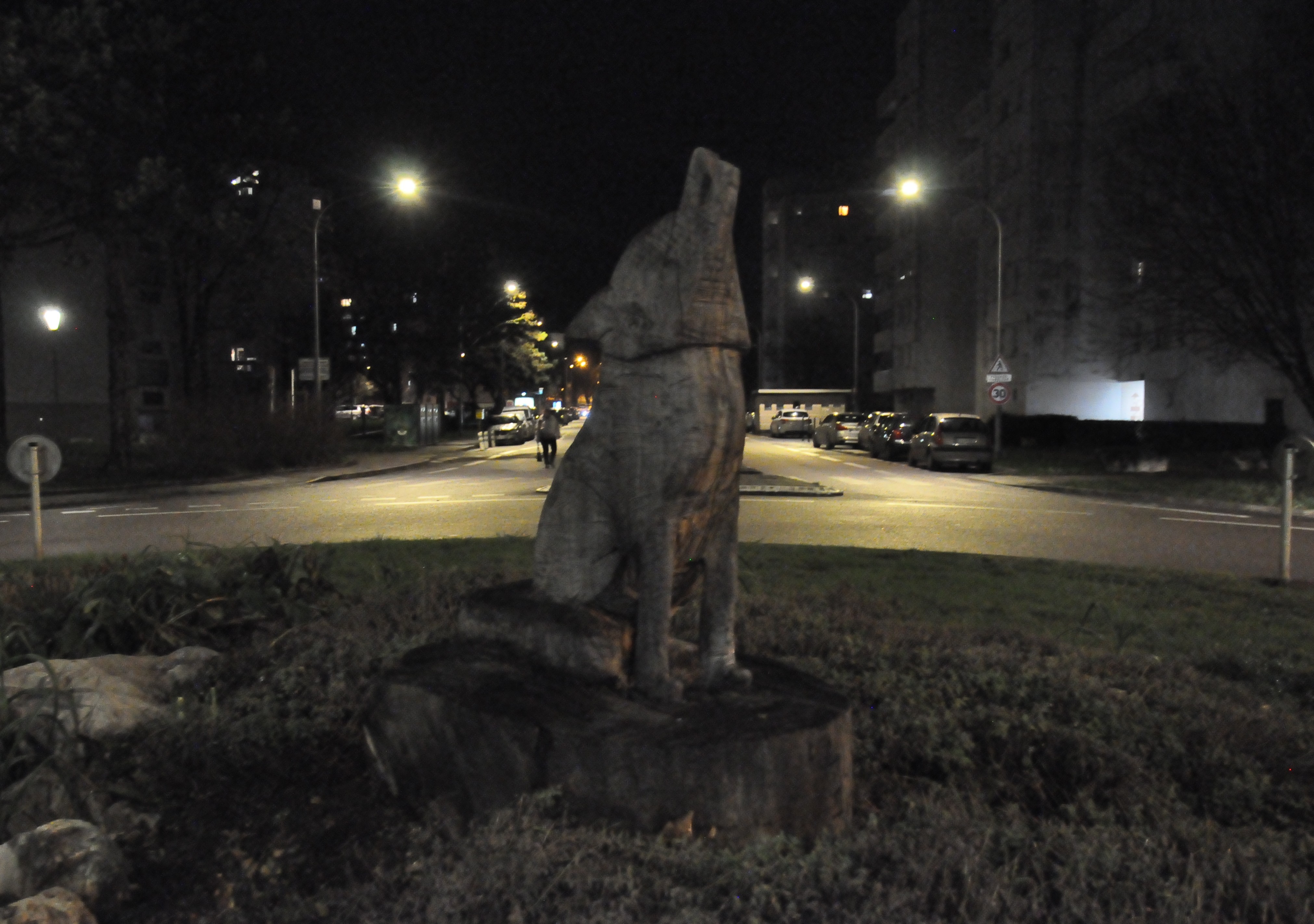 Sculpture en bois d'un loup en train de hurler, posée sur une souche d'arbre, sur un rond-point. L'ensemble est relativement sombre, malgré les éclairages de la route.