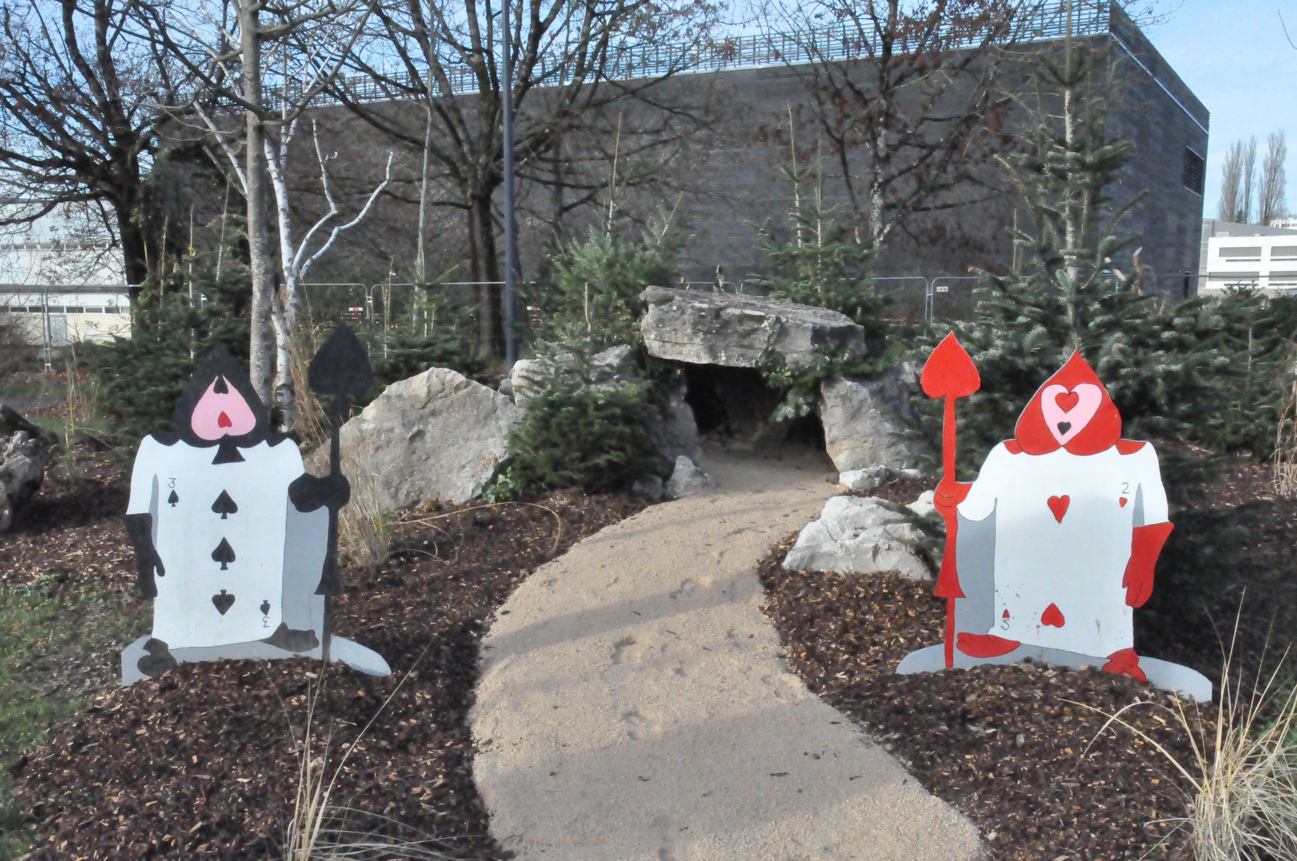 Photo d'une petite grotte aménagée sous des pierres, entourée de sapins, gardée par deux effigies de soldats "carte à jouer" issus d'Alice au pays des merveilles.