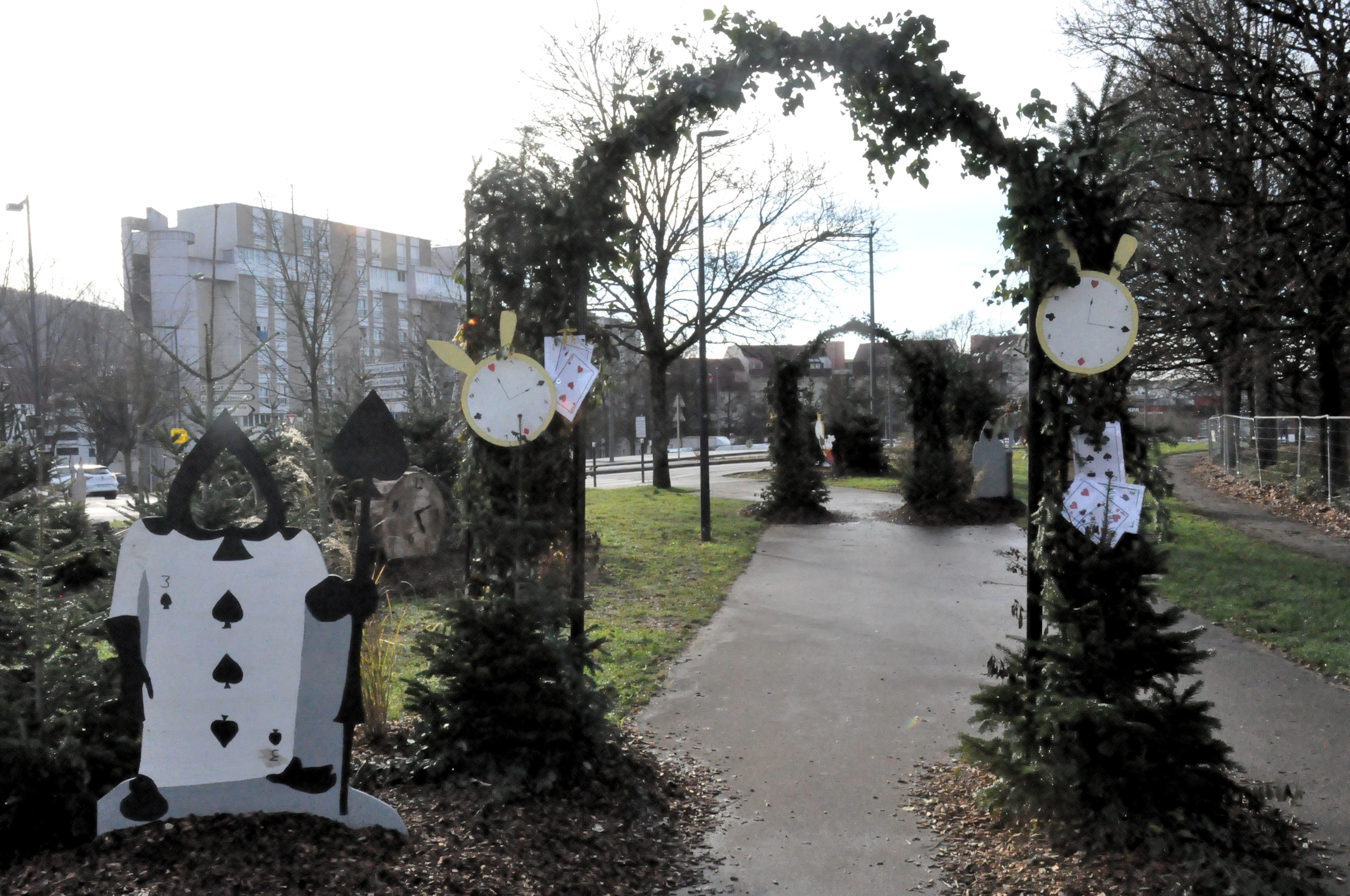 Dézoom de l'image précédente, où le panneau est en réalité une arche, agrémentée des cartes et des horloges de chaque côté, gardée par une carte à jouer 3 de pic. Plusieurs arches se succèdent autour d'un chemin sur le trottoir.