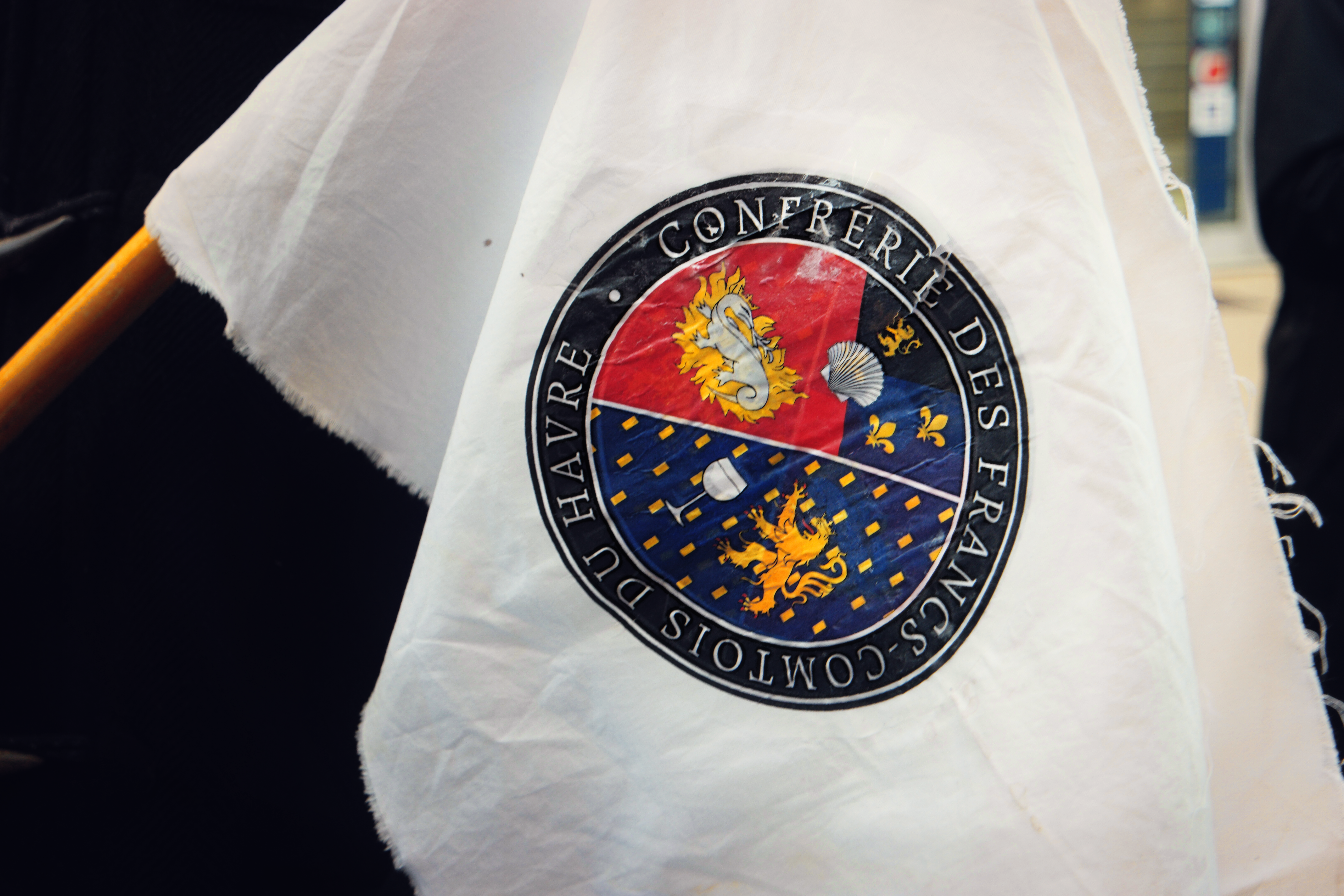 Un drapeau blanc floqué d'un cercle divisé en deux, une partie représentatnt un lion doré sur un fond bleu à côté d'un verre de vin, l'autre un dragon dans ses flammes sur fond rouge, à côté d'un côté coquillage et de deux fleurs de lys et d'un lion sur fond bleu. Le pourtour est ainsi intitulé : confrérie es francs-comtois du Havre