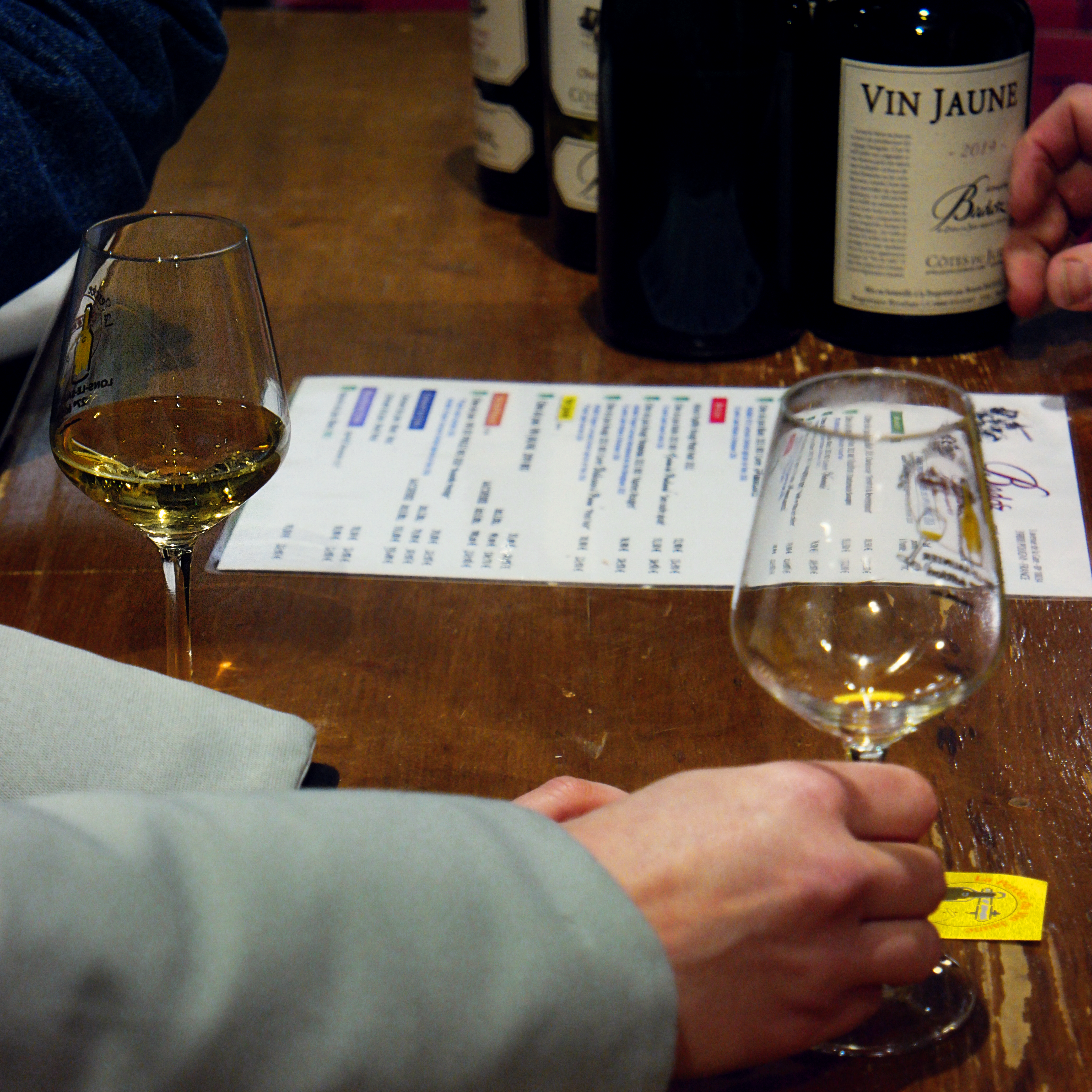 photo de mains tenant des verres de vin jaune sur un comptoir, avec une feuille de tarifs