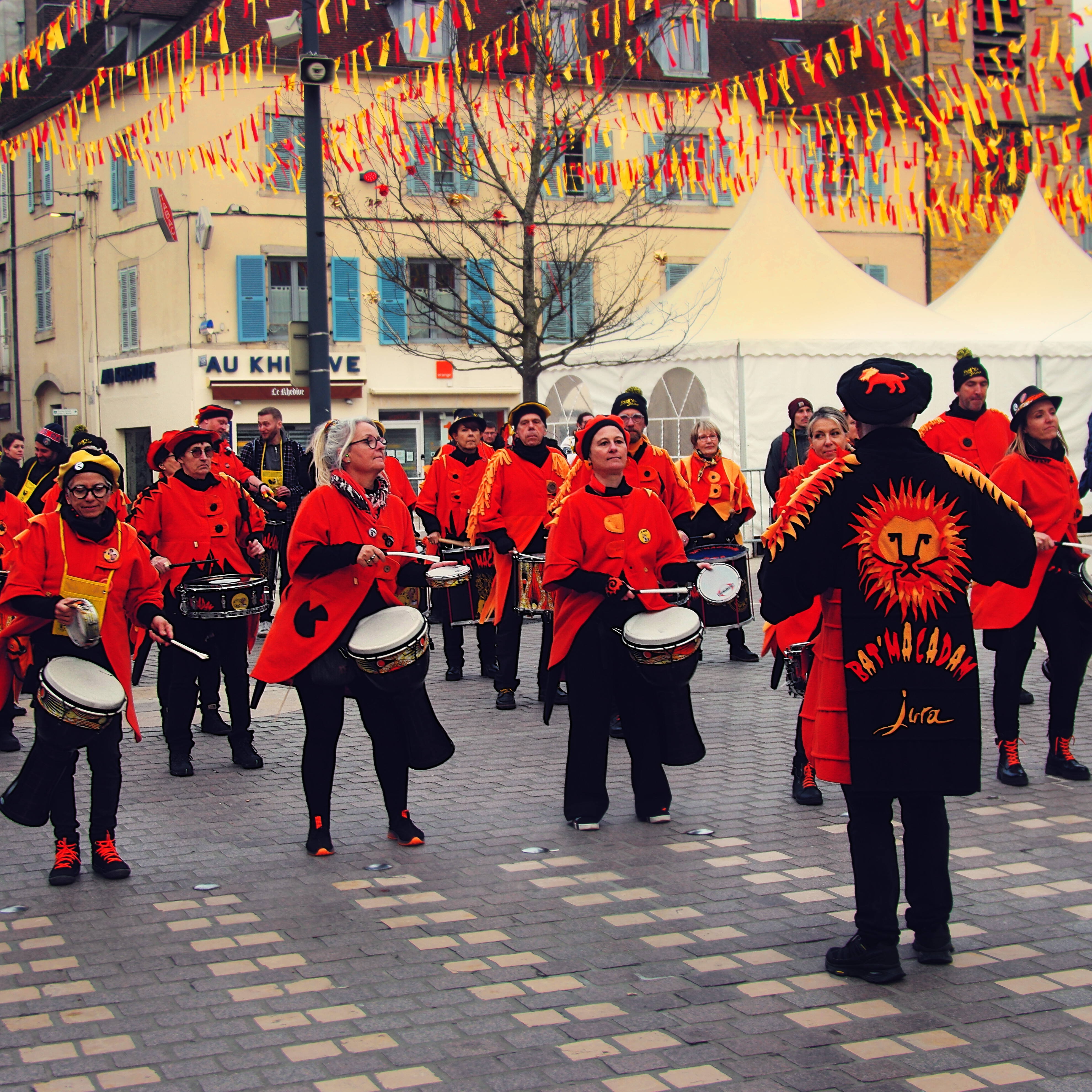 batucada du groupe "Bat-macadam", avec ses musicien·nes habillées de rouge et son chef d'orchestre portant des vêtements noirs floqués d'un lion rouge