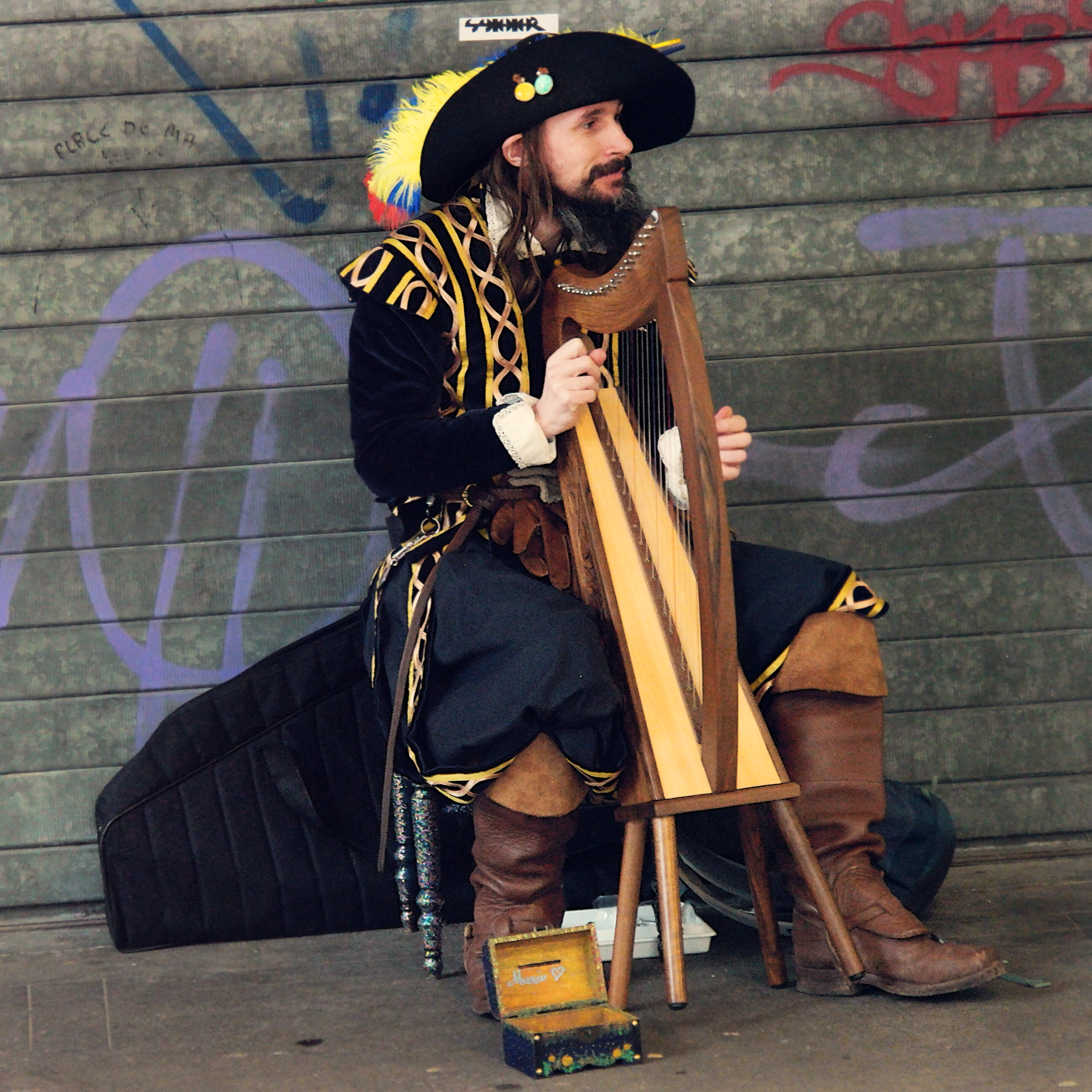 harpiste de rue jouant d'une petite harpe celtique en bois, assis devant un ridaeu de fer. Il est déguisé en mousquetaire.