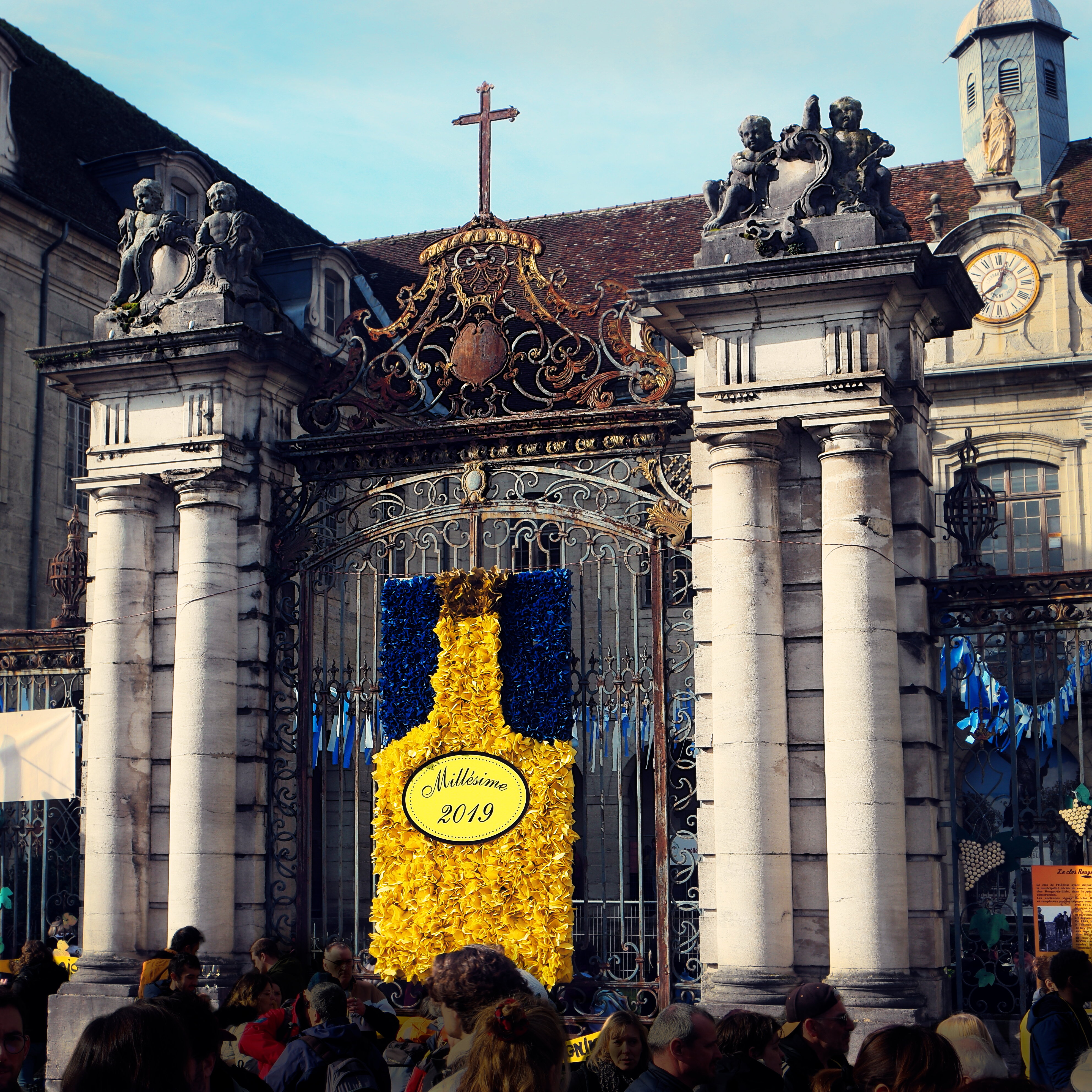 photo du grand portail de l'hôpital, avec ses colonnes et sa grille en métal sur laquelle a été appliquée une décoration de l'événement, une bouteille de vin jaune stylisée estampillée "millésime 2019"