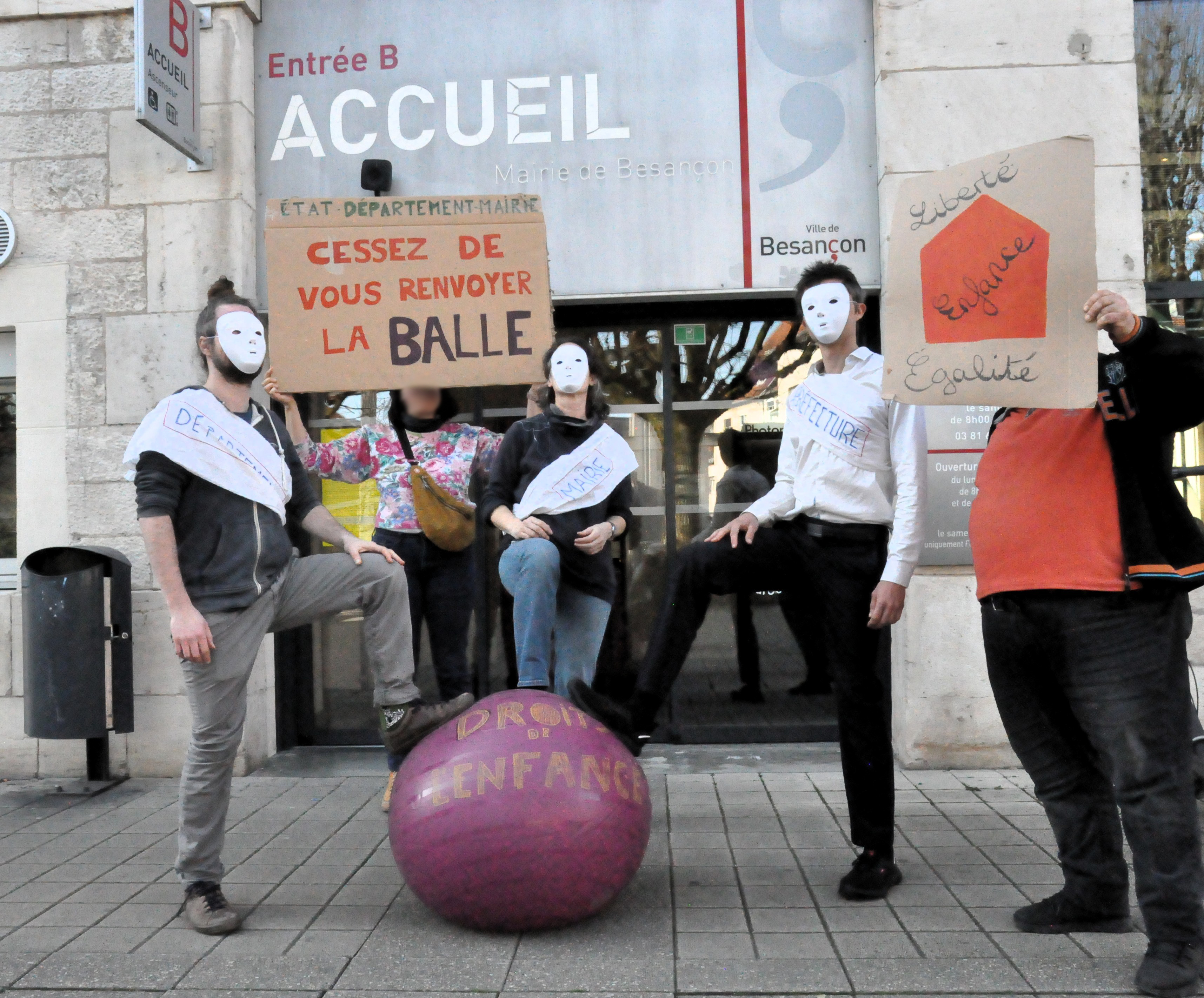 Les trois personnes masquées en blanc posent à nouveau le pied sur le ballon de gymnastique "droits de l'enfance", avec la pancarte "état, département, mairie, cessez de vous renvoyer la balle", cette fois devant l'entrée de la mairie. Une autre personne tient une pancarte "liberté, égalité, enfance"