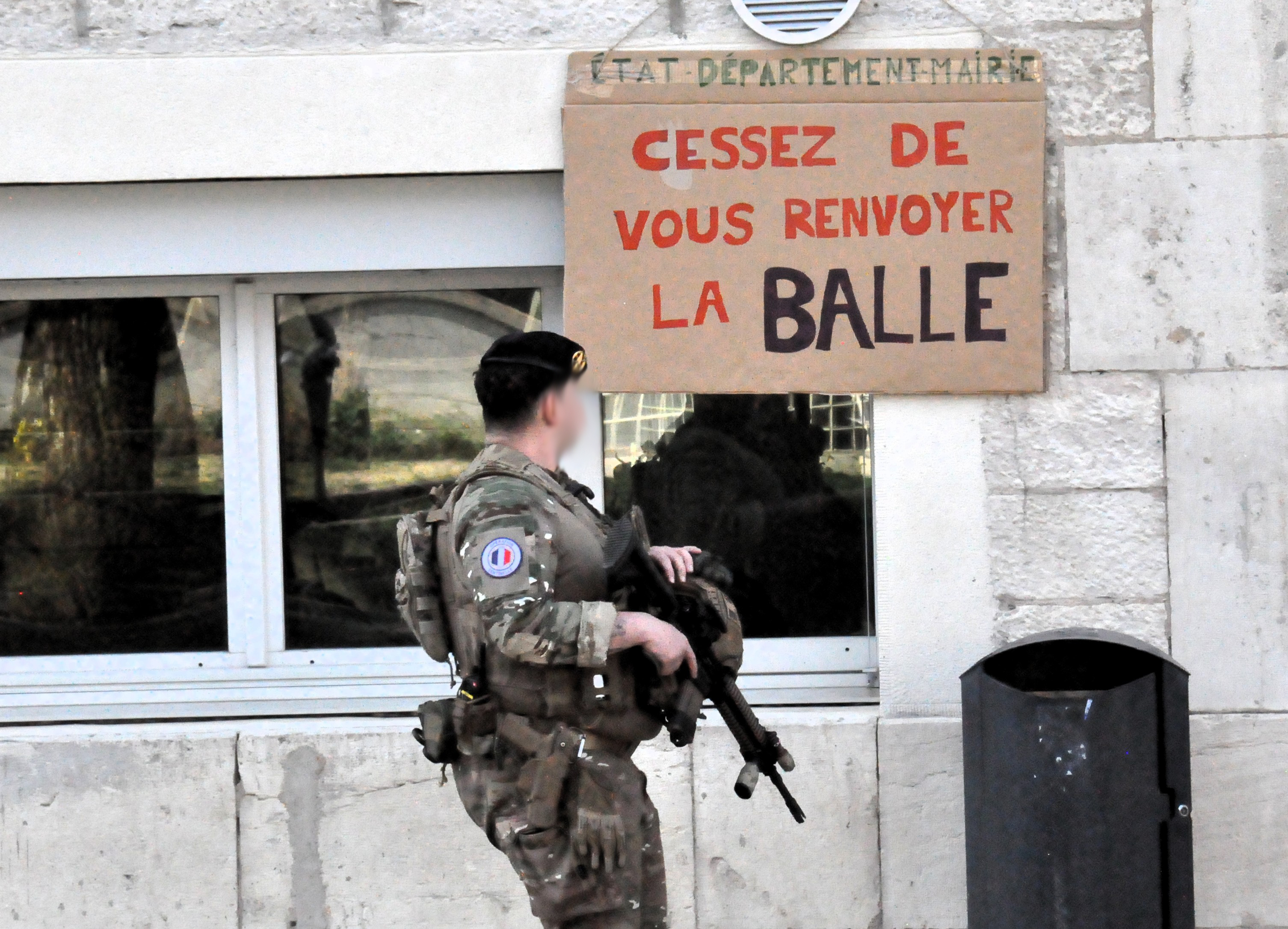 Un militaire armé en train de lire la pancarte "état, département, mairie, cessez de vous renvoyer la balle" accrochée devant la mairie.