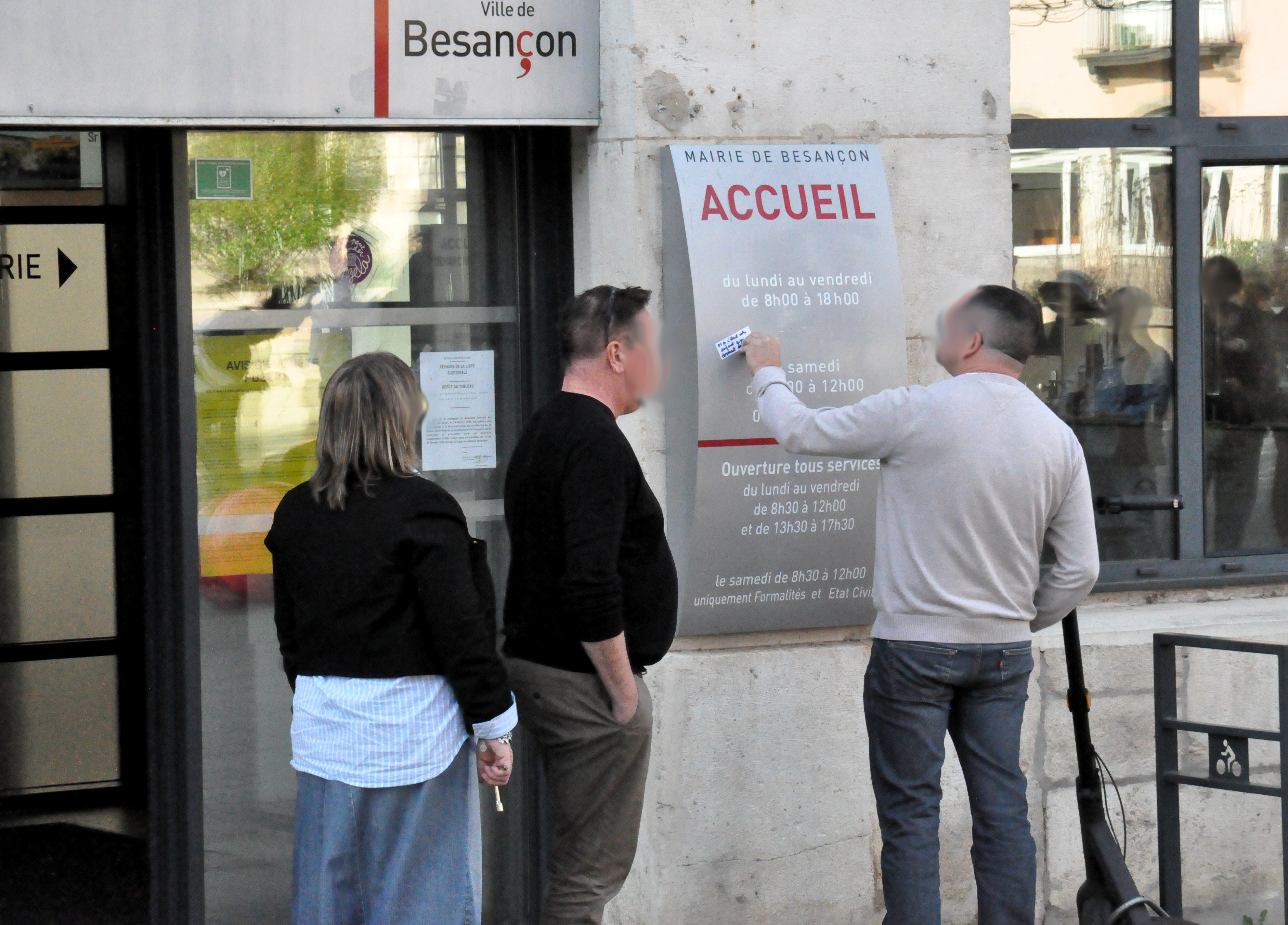 Trois personnes devant le panneau d'accueil de la mairie, dont une visiblement en train d'essayer d'enlever le sticker "et si c'était votre enfant qui dormait dehors ?" récemment collé.
