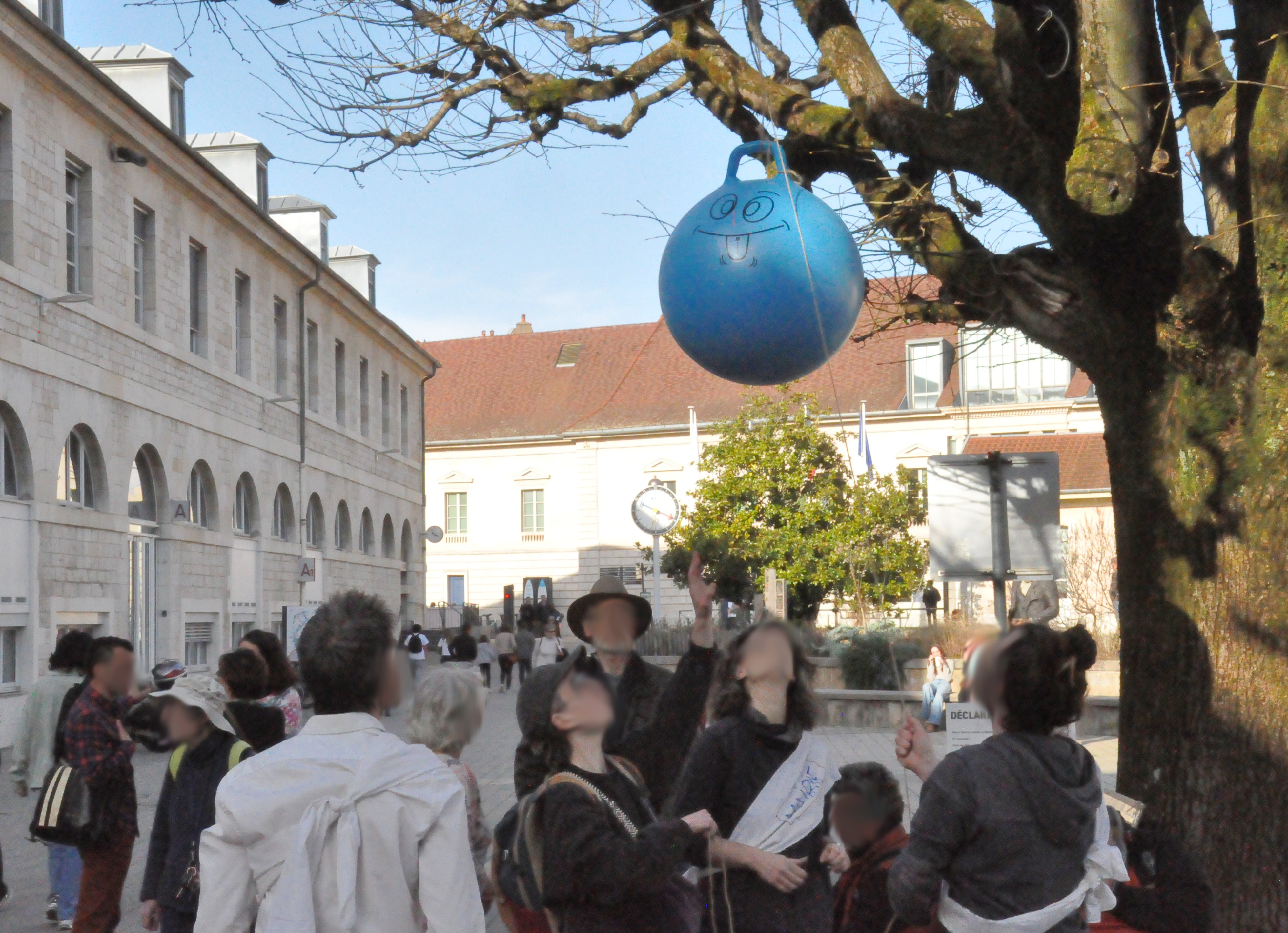 Le groupe de personne est en train de hisser un ballon de gymnastique bleu dans un arbre de l'esplanade des droits de l'homme, devant la mairie.