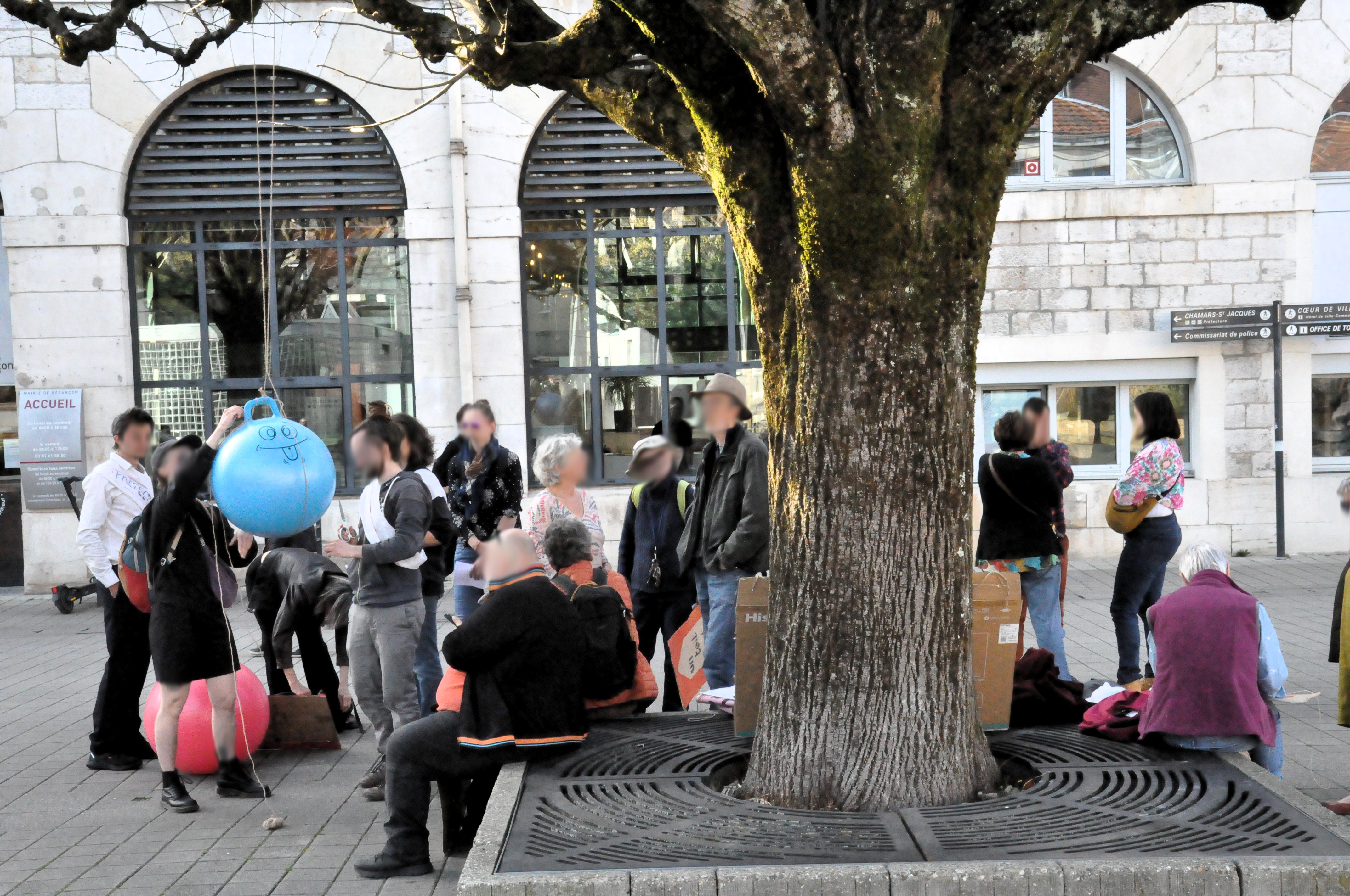 Le groupe de personne est en train de hisser un ballon de gymnastique bleu dans un arbre de l'esplanade des droits de l'homme, devant la mairie, sous un autre angle.