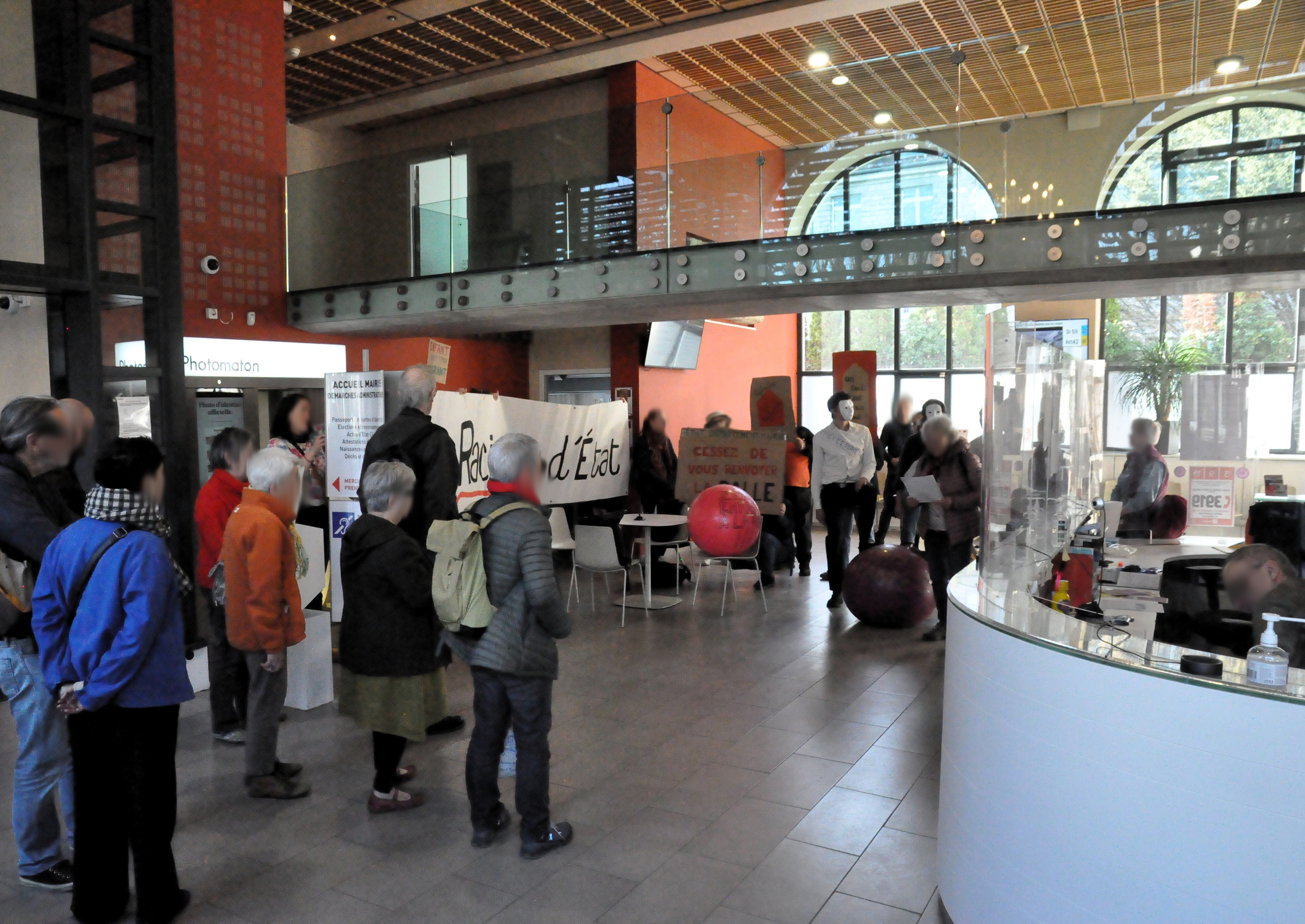 Les trois personnes masquées en blanc posent à nouveau le pied sur le ballon de gymnastique "droits de l'enfance", avec la pancarte "état, département, mairie, cessez de vous renvoyer la balle", cette fois devant l'entrée de la mairie. Une autre personne tient une pancarte "liberté, égalité, enfance", sous un autre angle. On aperçoit aussi une pancarte "racisme d'état".