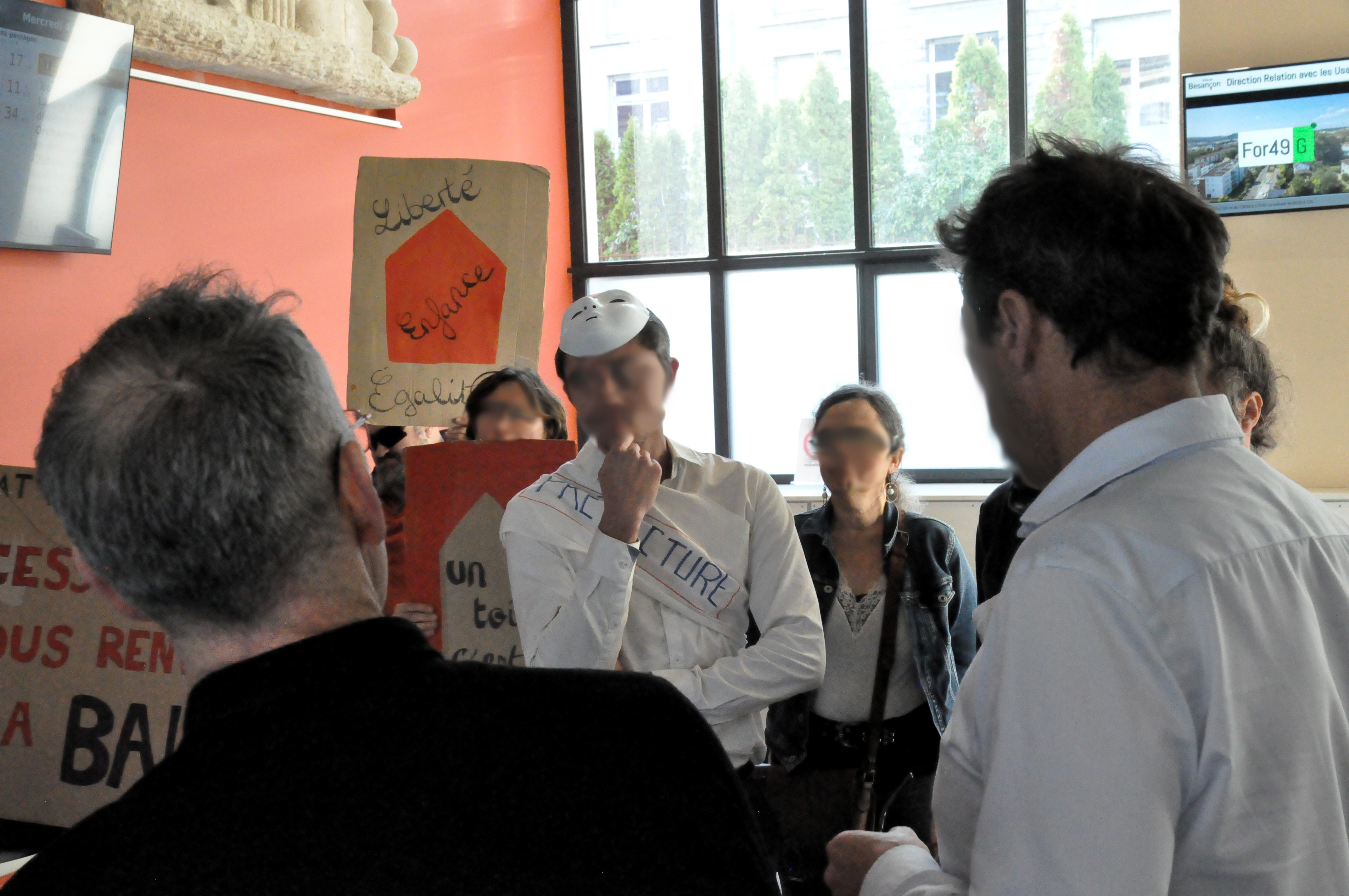 Une personne a retiré son masque blanc et se tient le menton dans un moment de réflexion. Derrière, on voit la pancarte "liberté, égalité, enfance", ainsi que certains bouts d'autres pancartes.