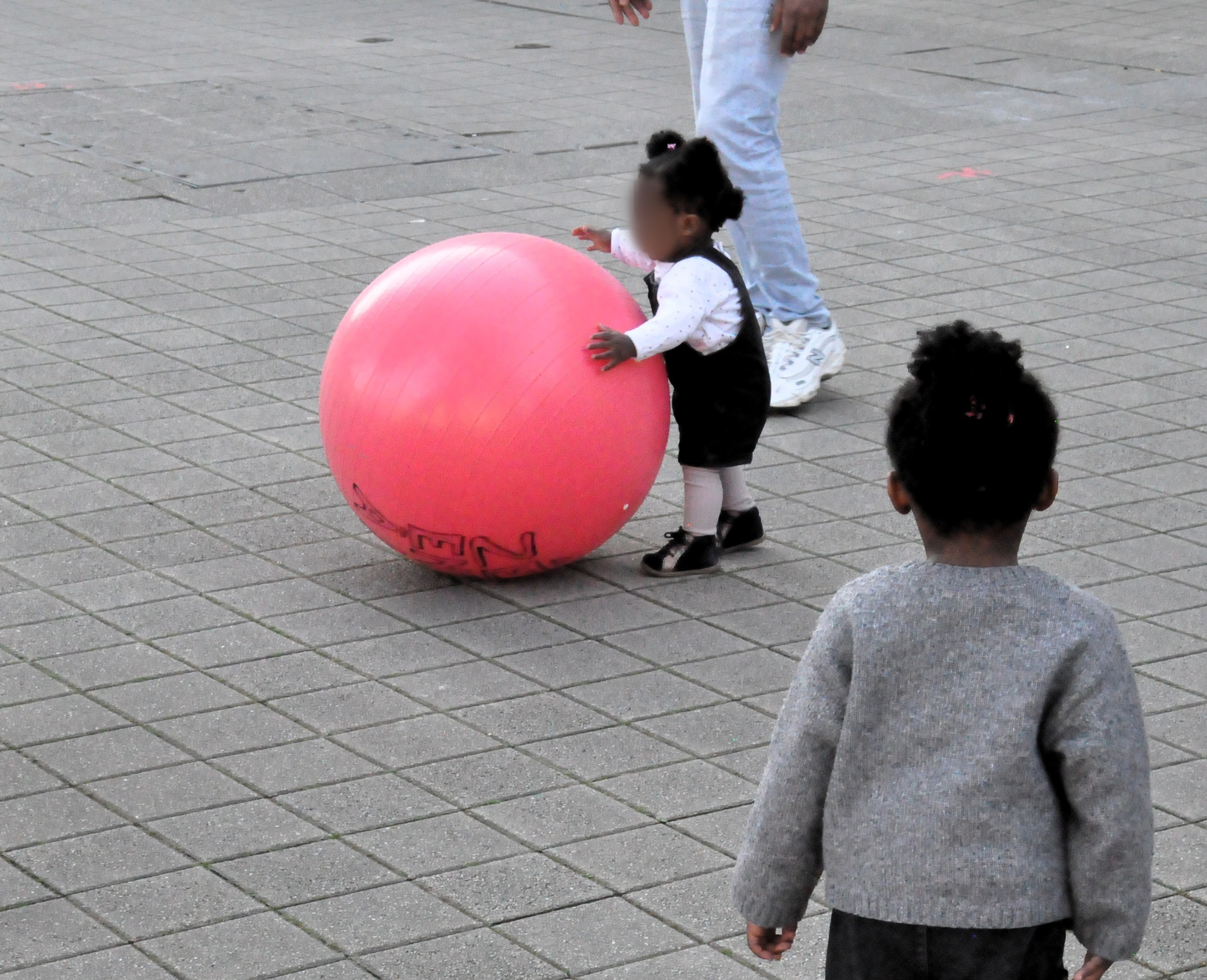 Une petite fille noire en train de jouer avec un des ballons de gymnastique sur le trottoir, sous la supervision de son père.