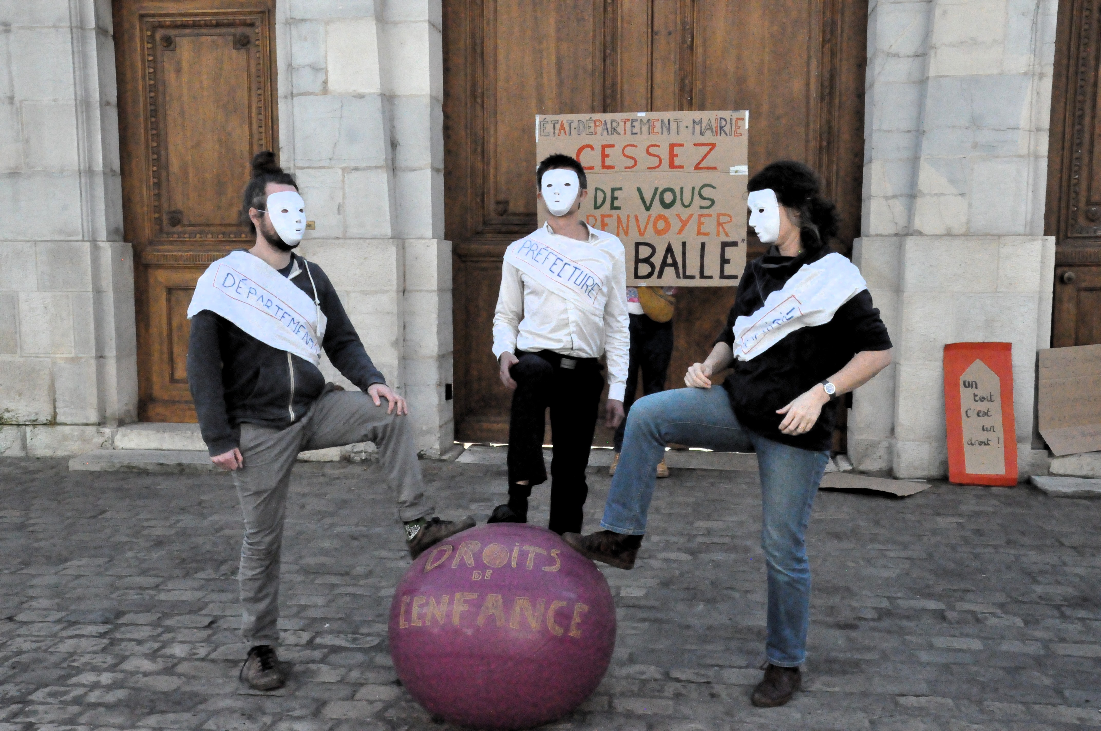 Trois personnes, portant chacune un masque blanc, mettent le pied sur un ballon de gymnastique sur lequel est écrit "droits de l'enfance", devant la préfecture. Derrière, une pancarte sur laquelle est inscrit "état, département, mairie, cessez de vous renvoyer la balle" est appuyée sur la porte.