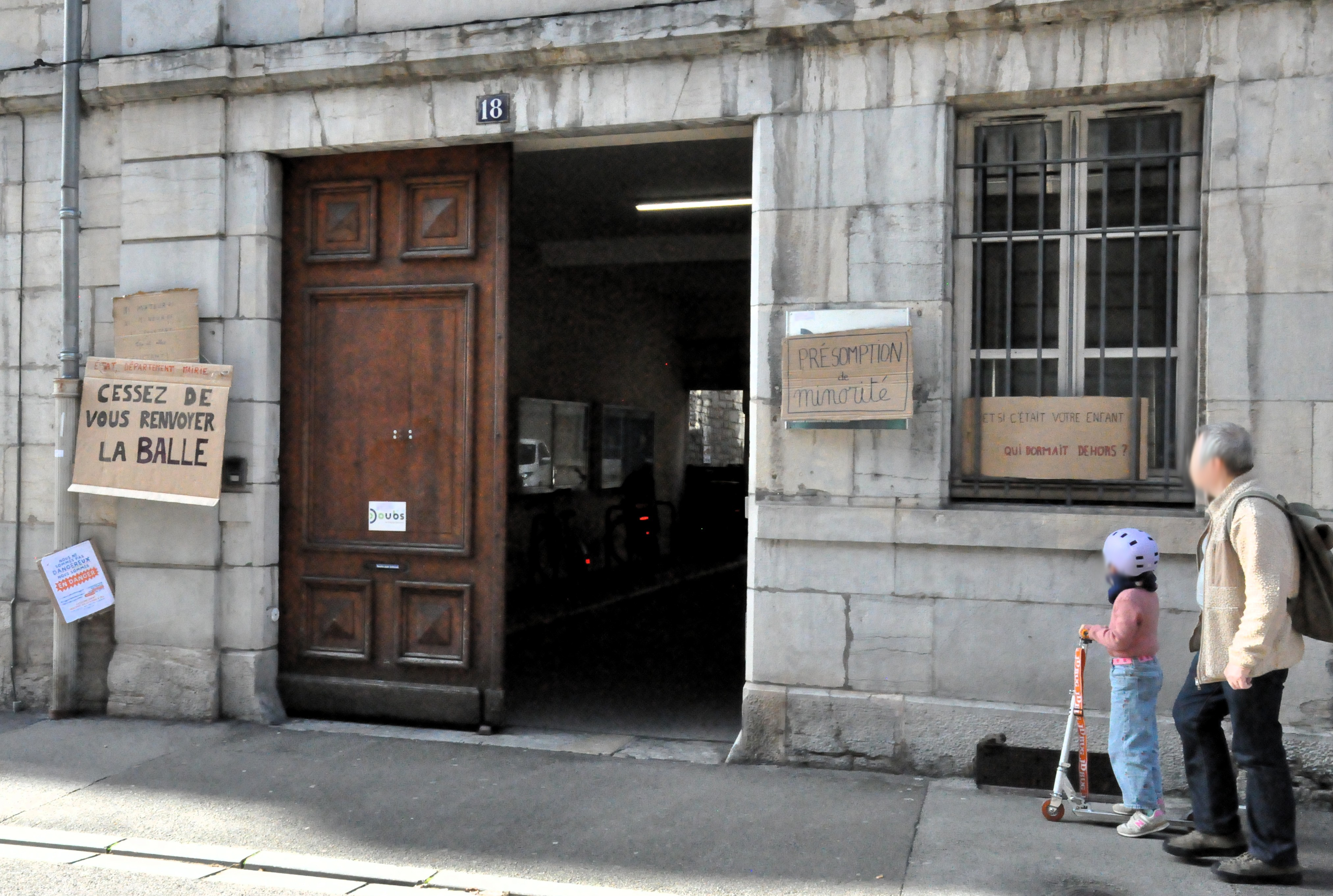 L'entrée des locaux du département du Doubs, avec la pancarte "état, département, mairie, cessez de vous renvoyer la balle" à gauche et "présomption de minorité" à droite. Encore à droite, une pancarte "et si c'était votre enfant qui dormait dehors ?" Une fillette à trottinette et son père passent à côté.