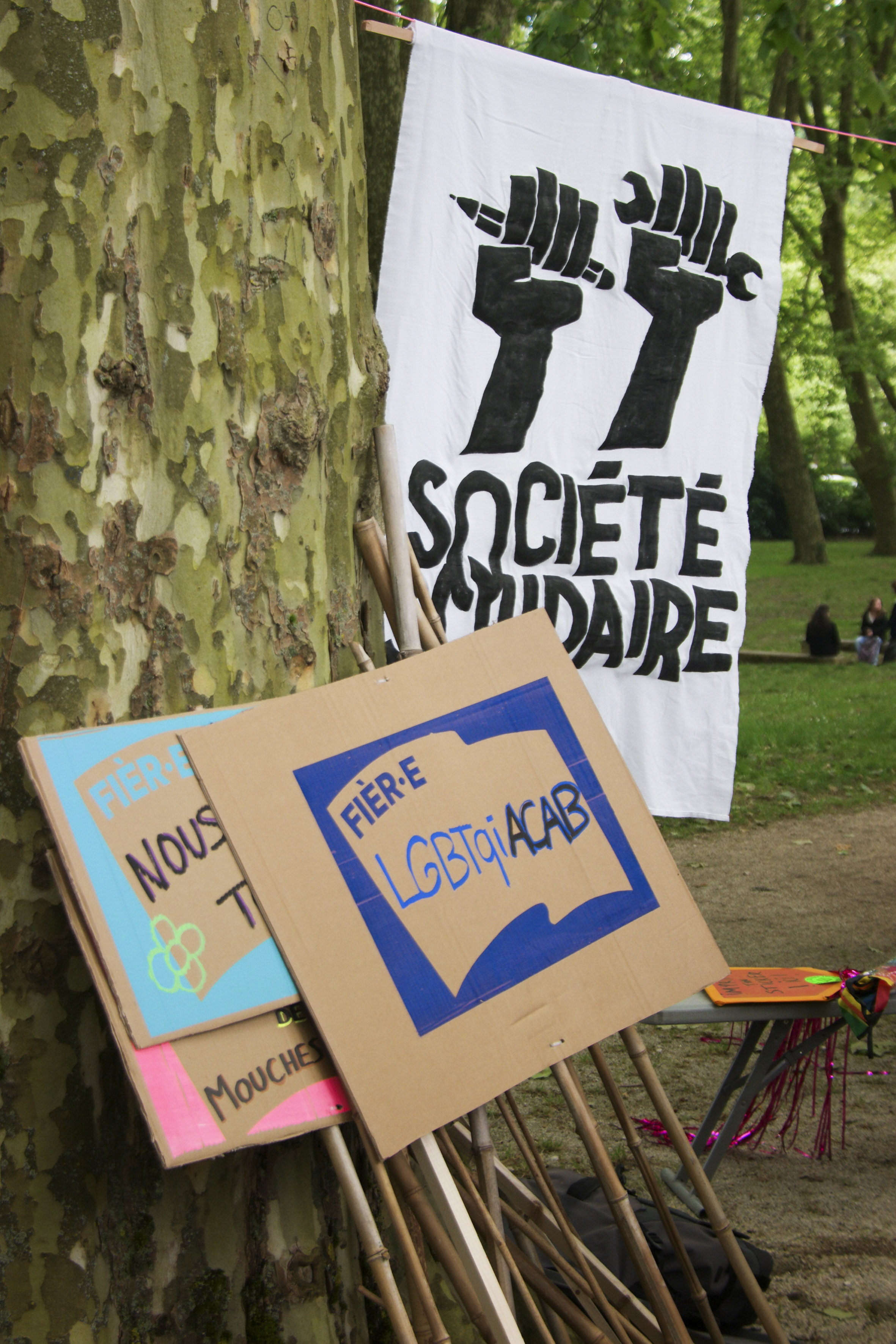 Un étendard Société solidaire avec deux poings levés, l'un tenant un crayon, l'autre une clé à molette, à côté d'une pancarte "fièr·e d'être LGBTQIACAB"