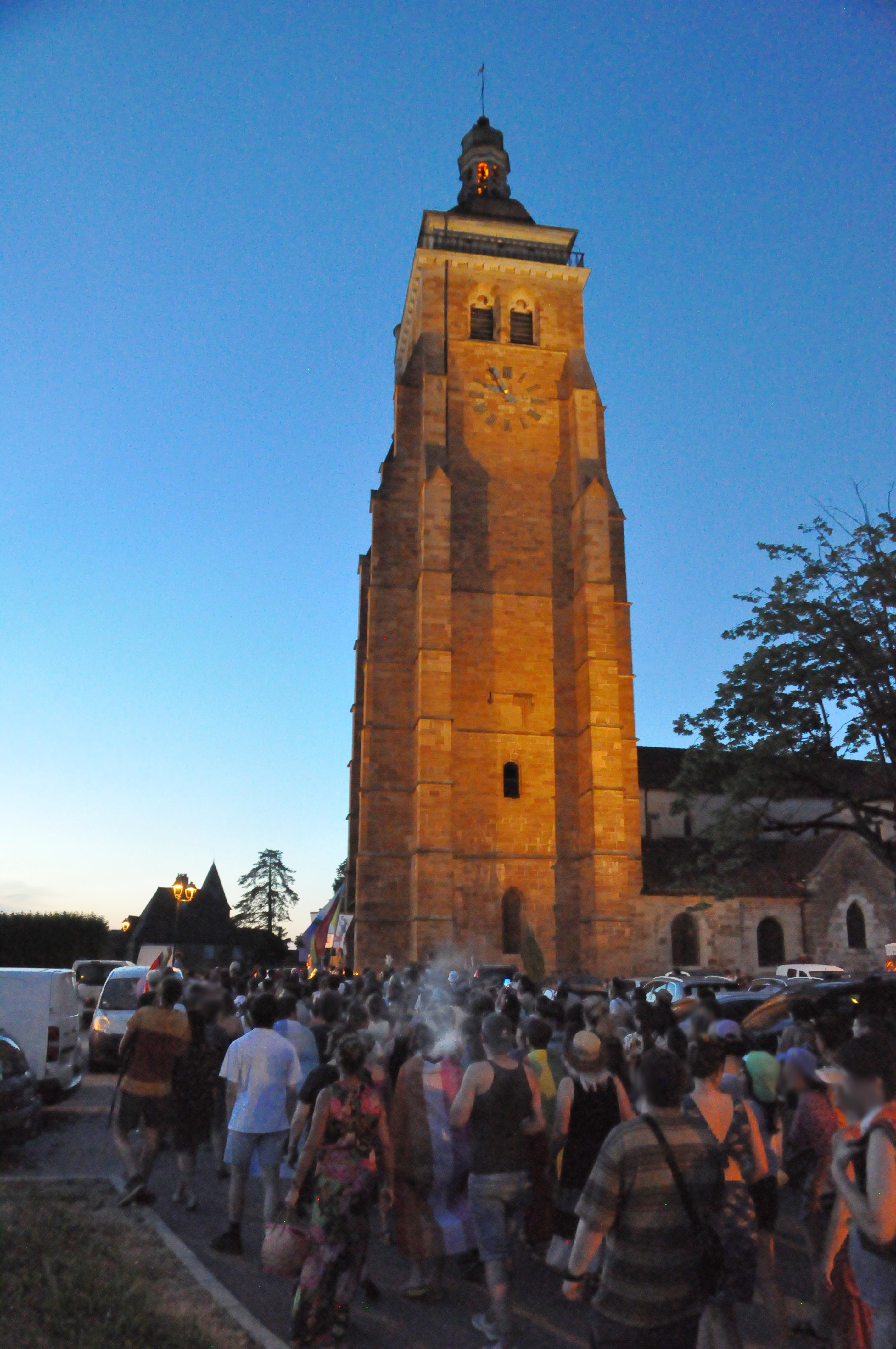 Défilé de la foule à la tombée du jour, au pied de l'église Saint-Just, éclairée par le bas