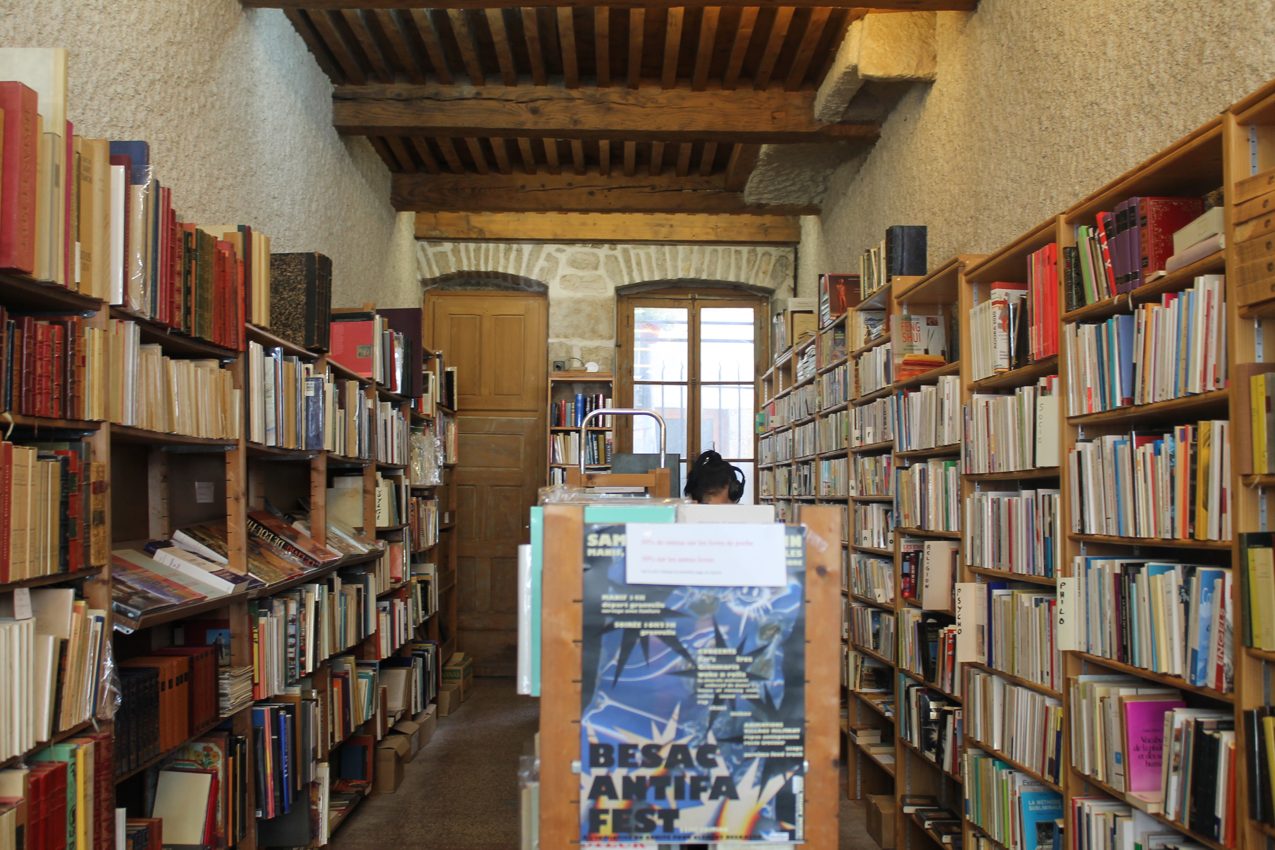 L'intérieur d'une librairie, au plafond de bois et aux murs de pierre enduits de crépit blanc. Entre les étagères en bois garnies de livre, l'espace est relativement exigu