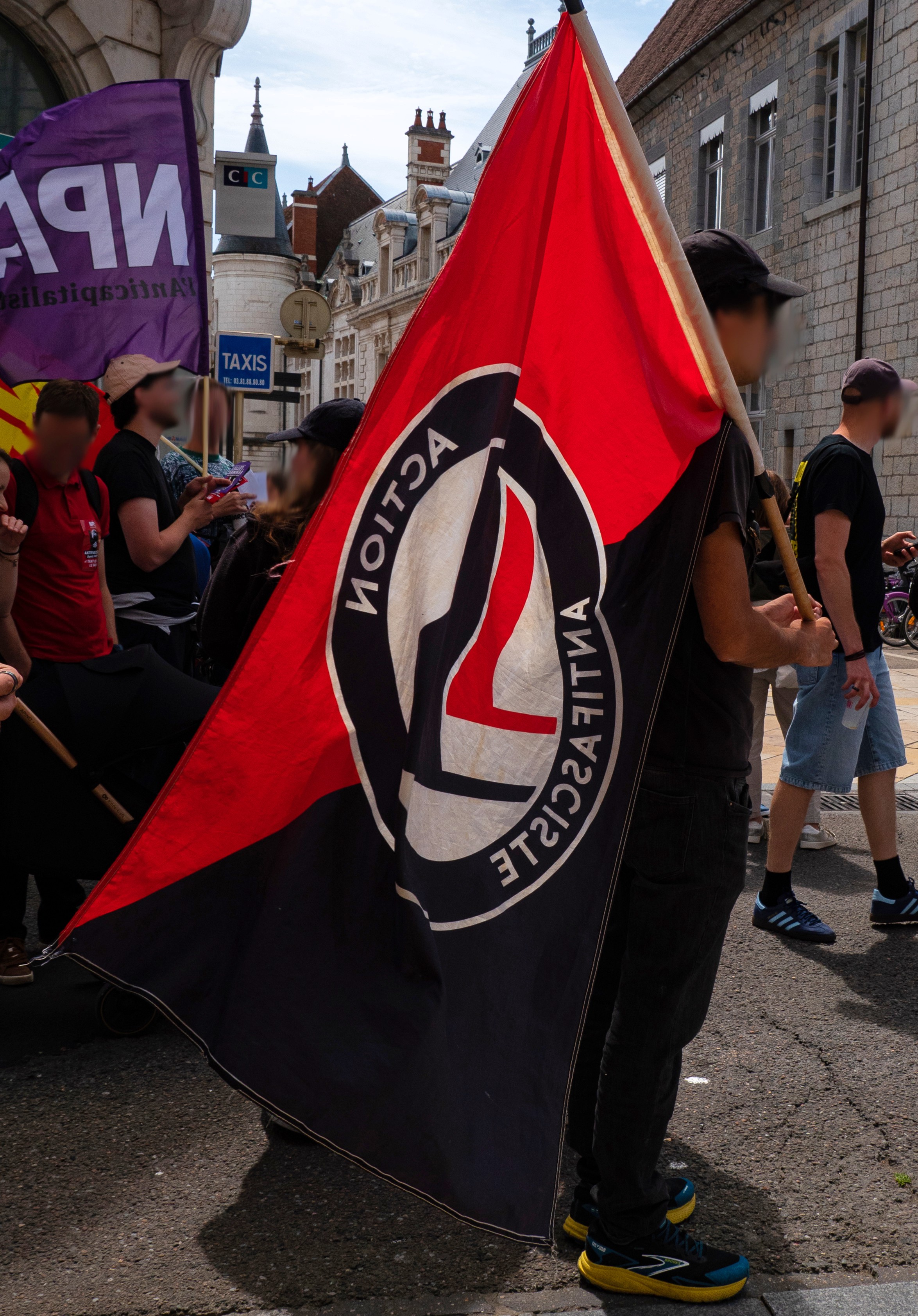 Une personne dans le cortège avec un drapeau Action Antifasciste rouge et noir aussi grand qu'elle. On aperçoit un drapeau du NPA juste derrière