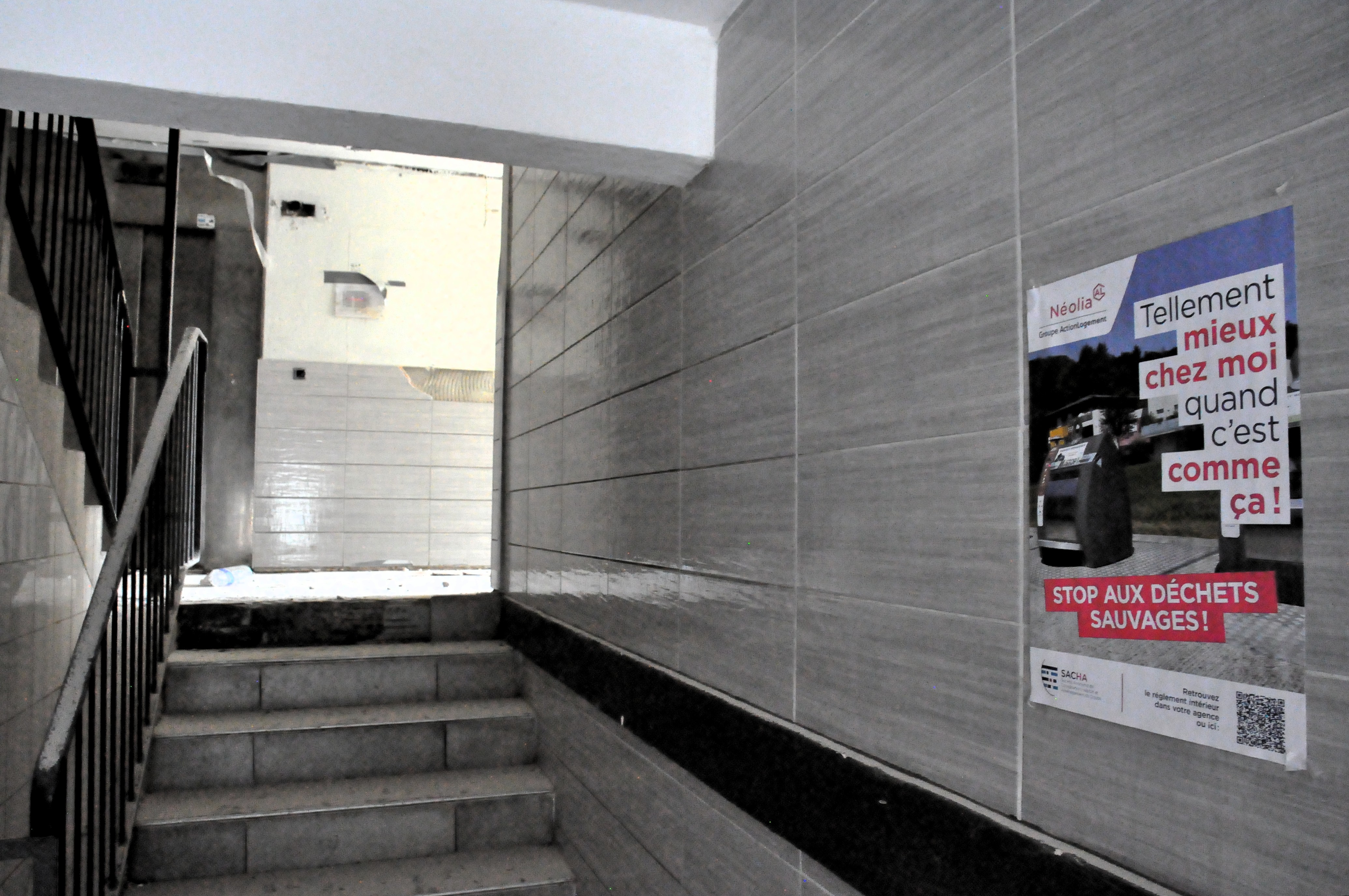 vue d'une cage d'escalier austère mal entretenue, en revêtement brillant gris, avec une affiche Néolia : tellement mieux comme ça quand c'est chez moi, appelant à stopper les déchets sauvages.