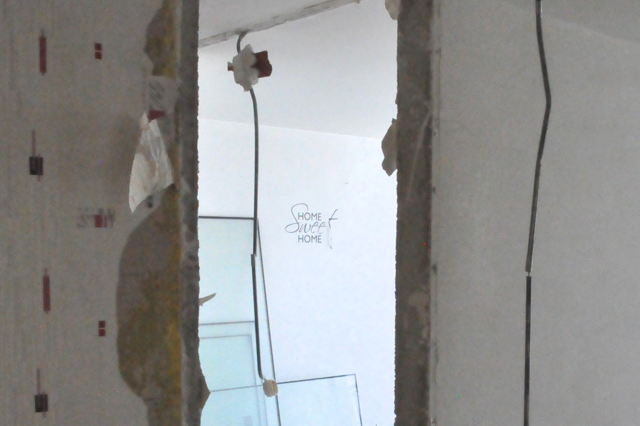une trouée dans un mur avec des fils électriques qui pendant. Sur le mur du fond, l'inscription Home Sweet Home