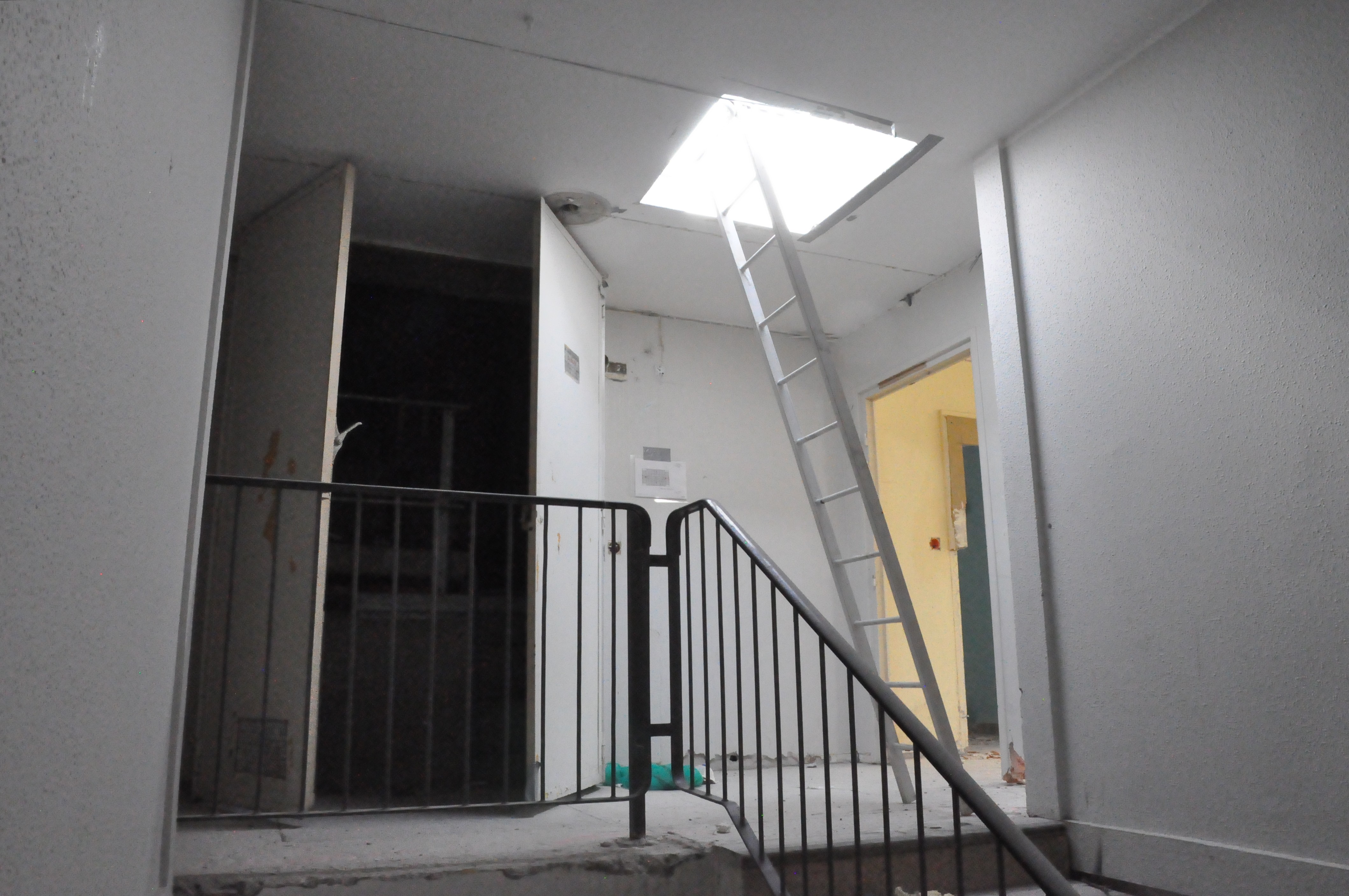 une cage d'escalier délabrée, avec un escabeau permettant l'accès à une trappe faisant puits de lumière au plafond