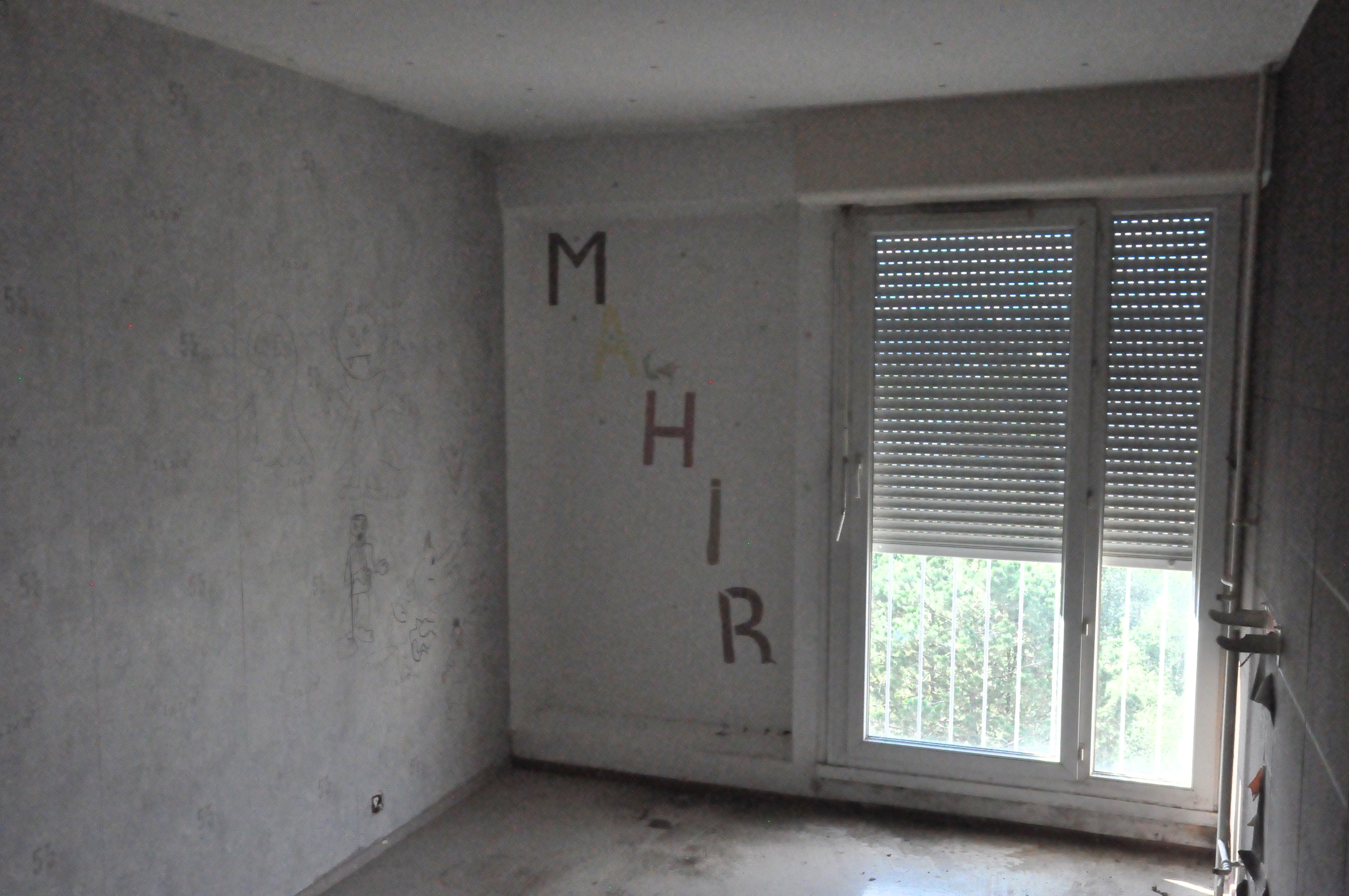 l'intérieur d'une chambre vide et délabrée, peu éclairée par la fenêtre semi ouverte, avec les lettres M A H I R inscrites en diagonales