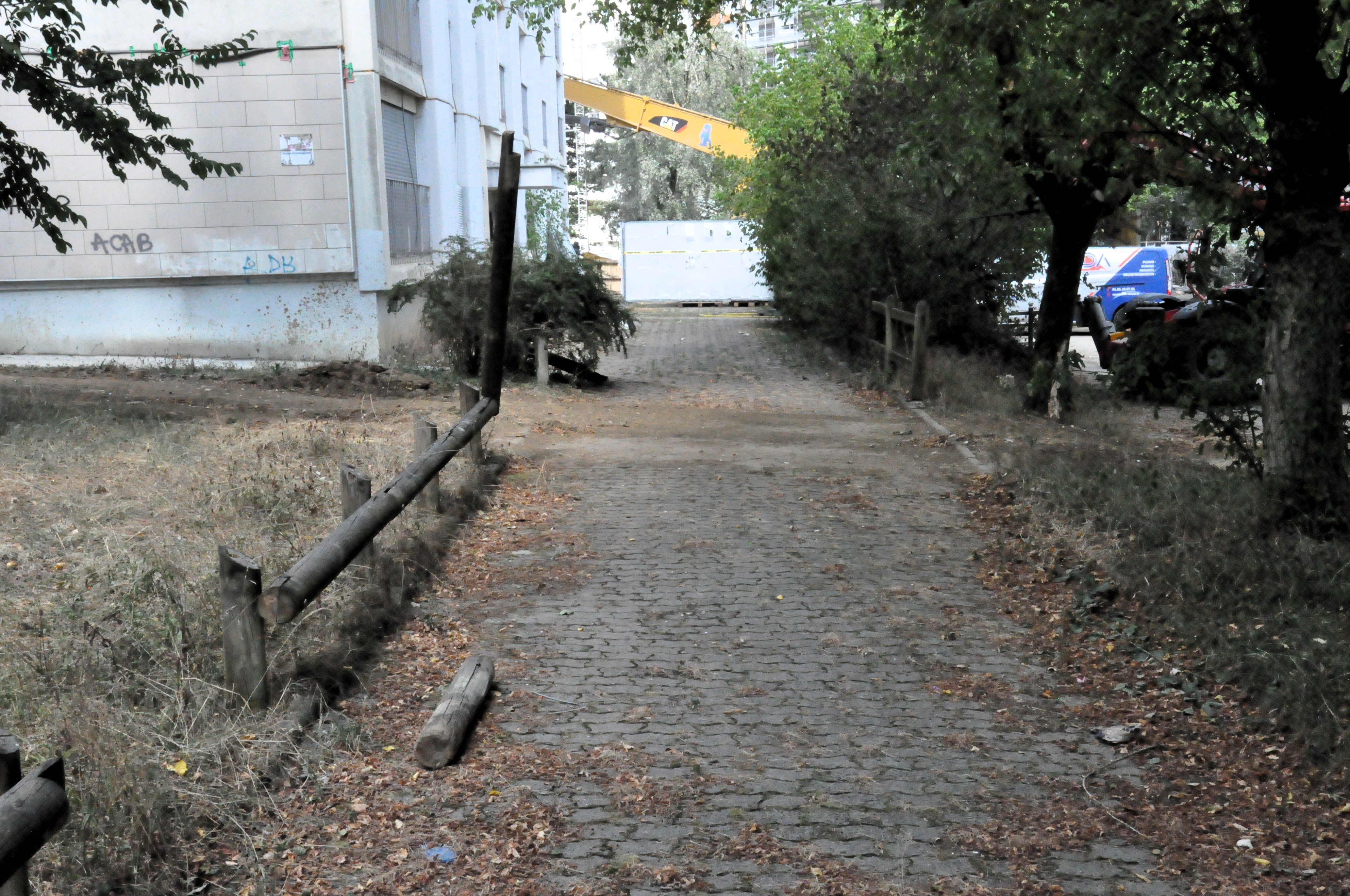 une allée de pavés autobloquants laissée sans entretien, avec un rondin de bois tombé de la petite barrière à côté du pré