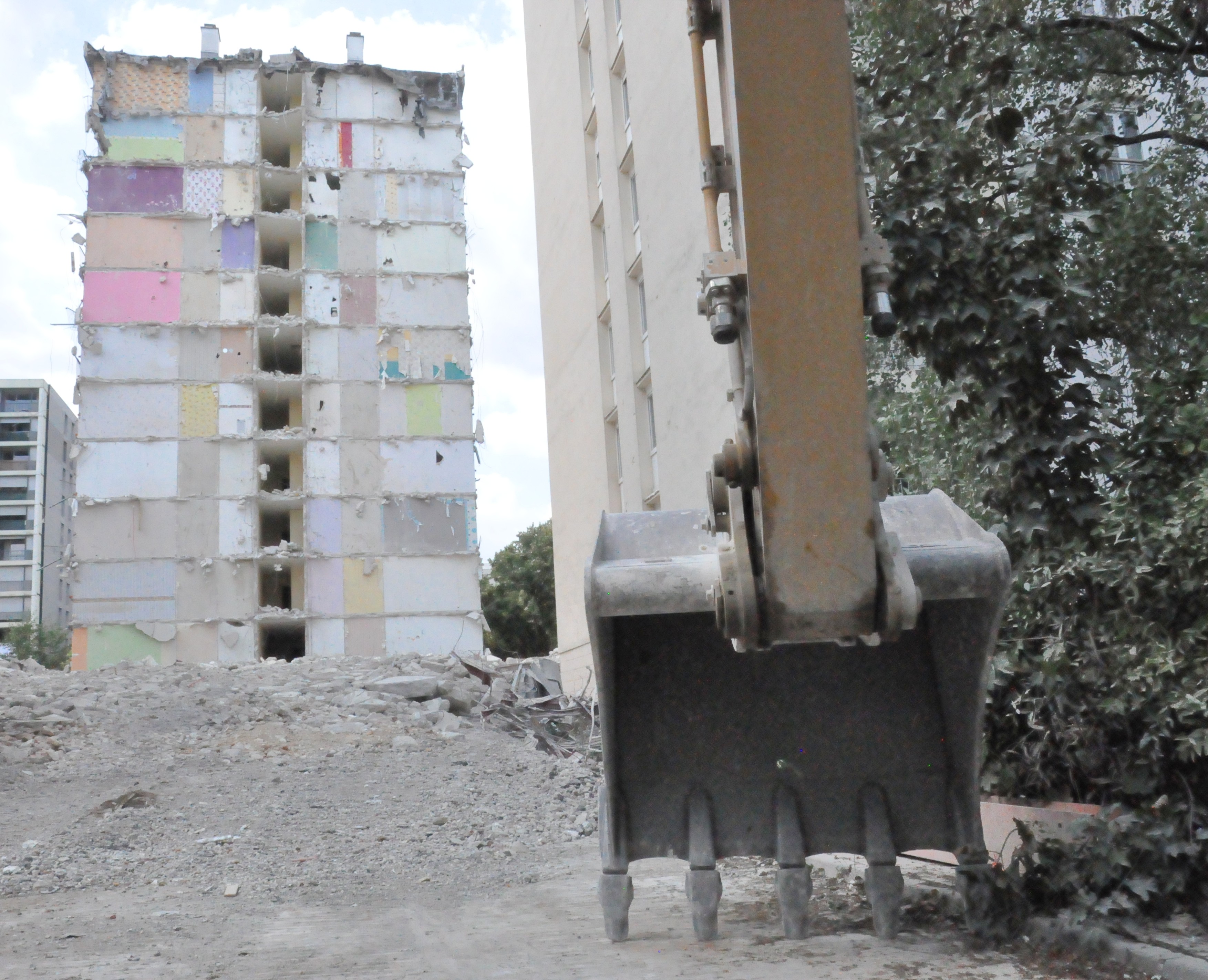le godet d'une pelleteuse face à un tas de gravats, avec un immeuble abandonné en fond, à la façade arrachée
