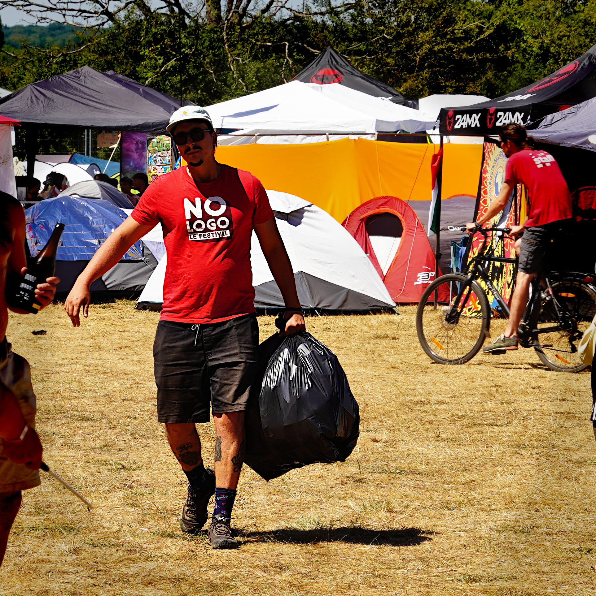 personne en t-shirt rouge No Logo festival qui transporte du matériel, sur fond de tentes installées sur l'herbe sèche façon camping 