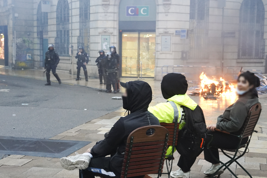 Des manifestant·e·s assis sur des chaises sur la place du 8 septembre 1945, en fond des policiers et une barricade en feu